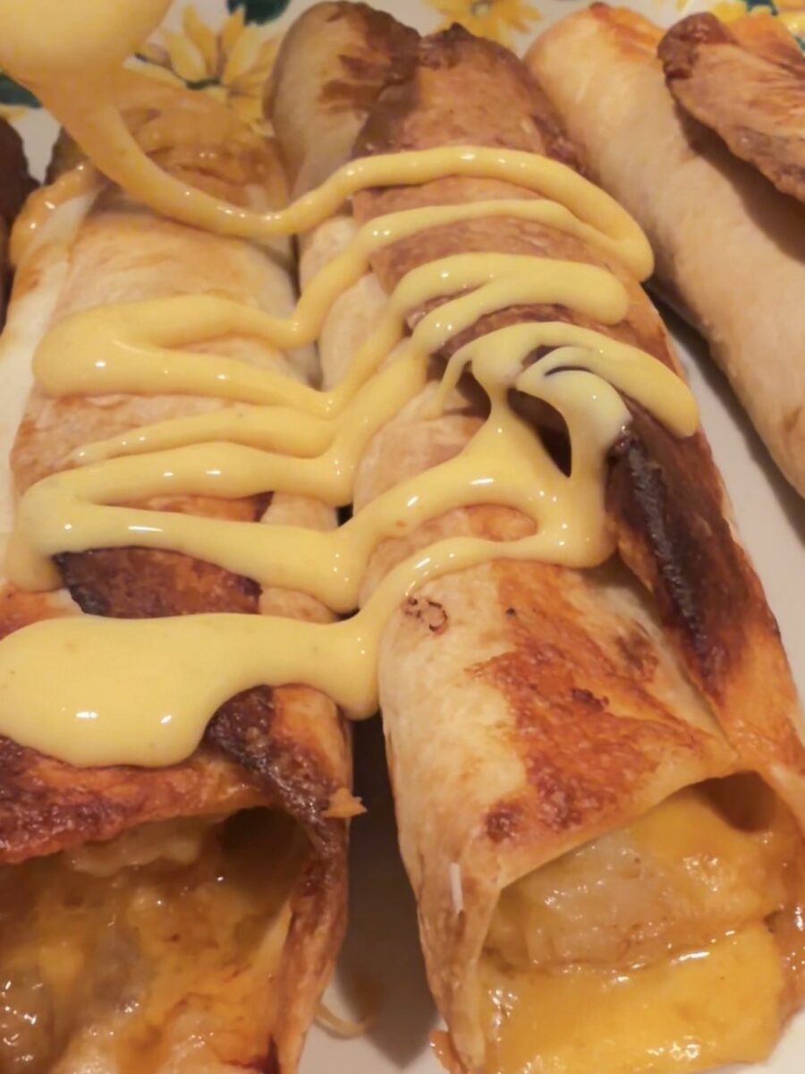 Three baked enchiladas topped with melted cheese are being drizzled with a creamy yellow sauce, served on a plate with a floral border.