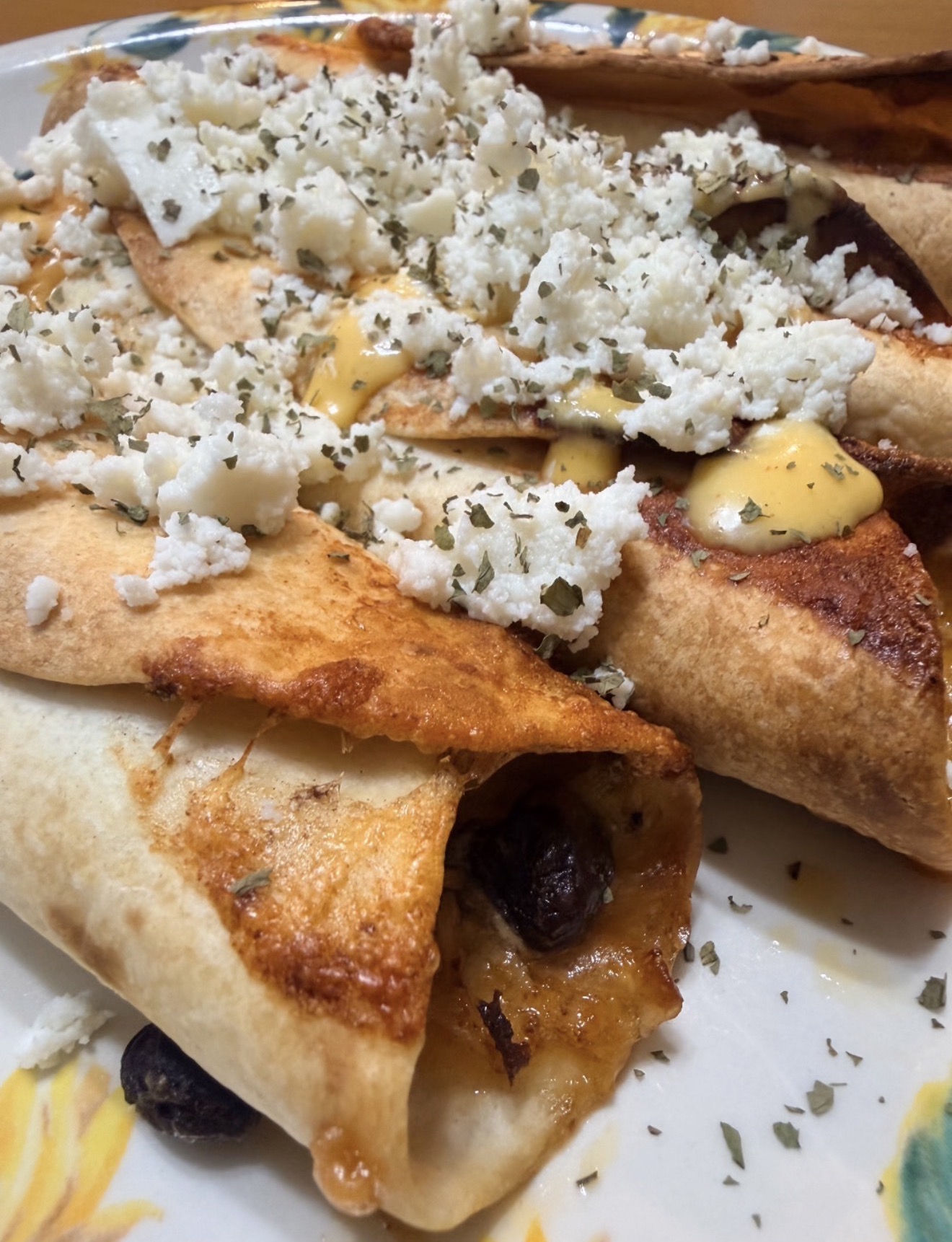 Close-up of crispy rolled tortillas filled with melted cheese and beans, topped with crumbled cheese, a drizzle of yellow sauce, and sprinkled herbs on a decorative plate.