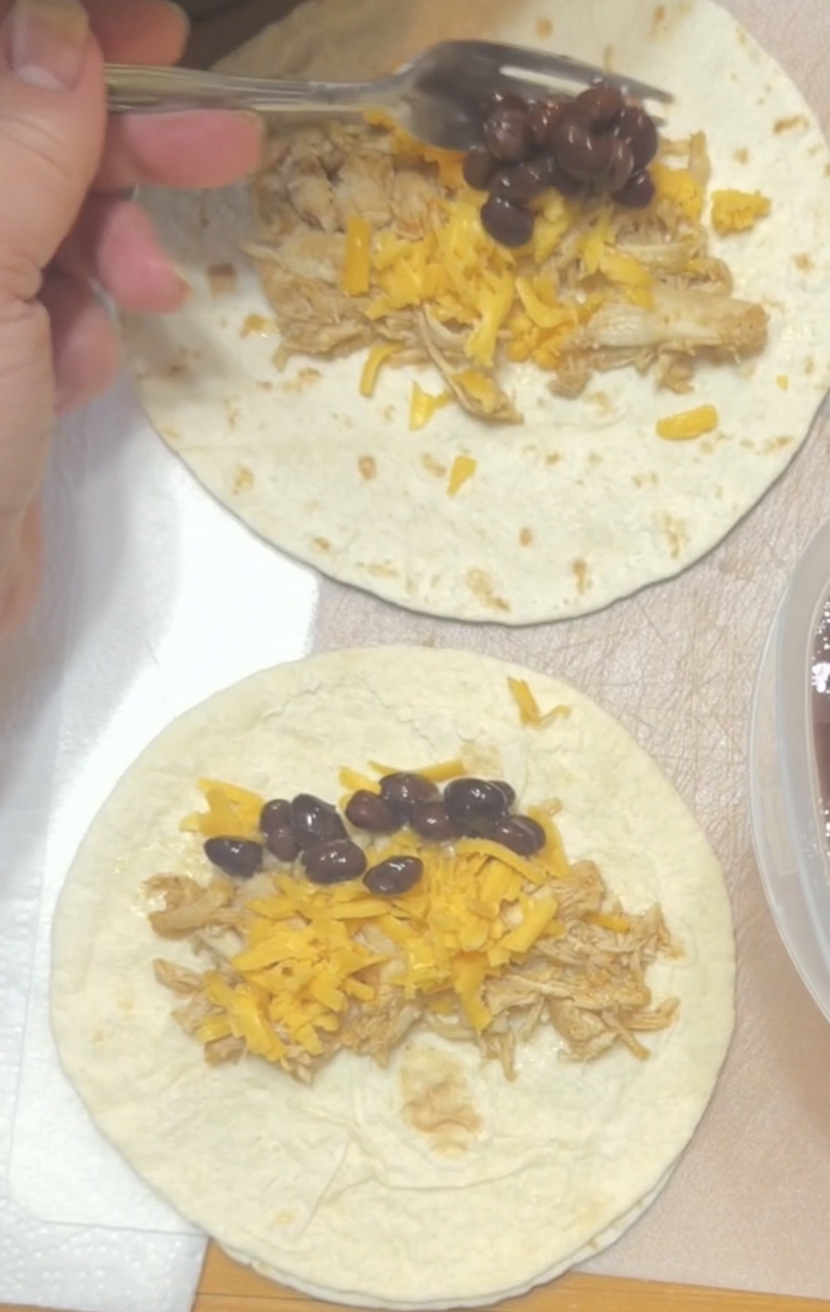 Two soft tortillas topped with shredded chicken, grated cheddar cheese, and black beans. A hand uses a spoon to add beans to one tortilla. The tortillas are on a countertop next to a container.