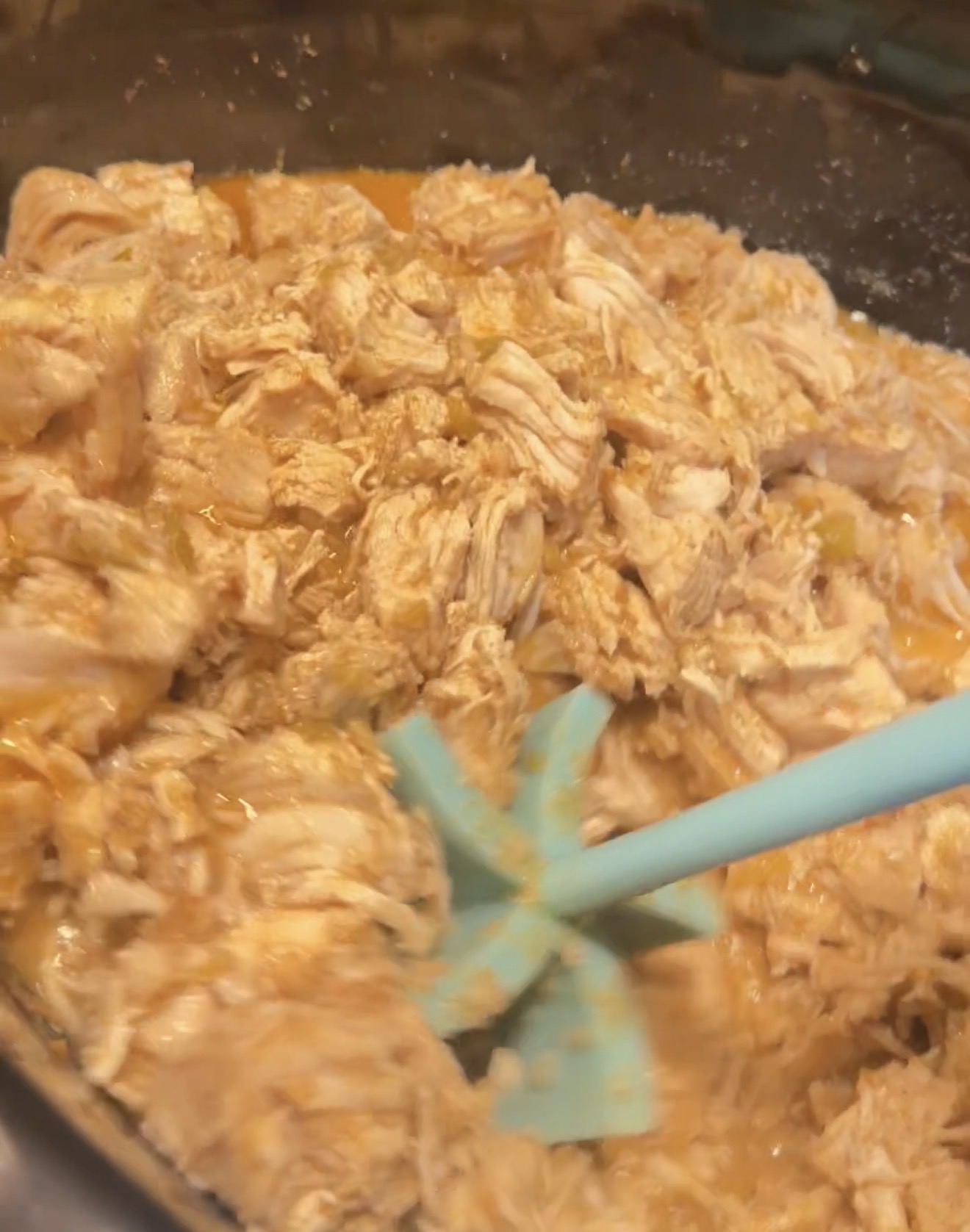 Shredded chicken mixed with sauce in a bowl, being stirred with a light blue spatula.