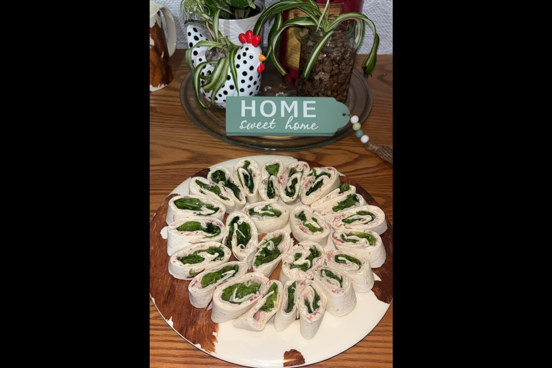 A plate of tortilla roll-up pinwheels filled with leafy greens and deli meat sits in front of a Home Sweet Home sign and ceramic chicken decor on a wooden table.