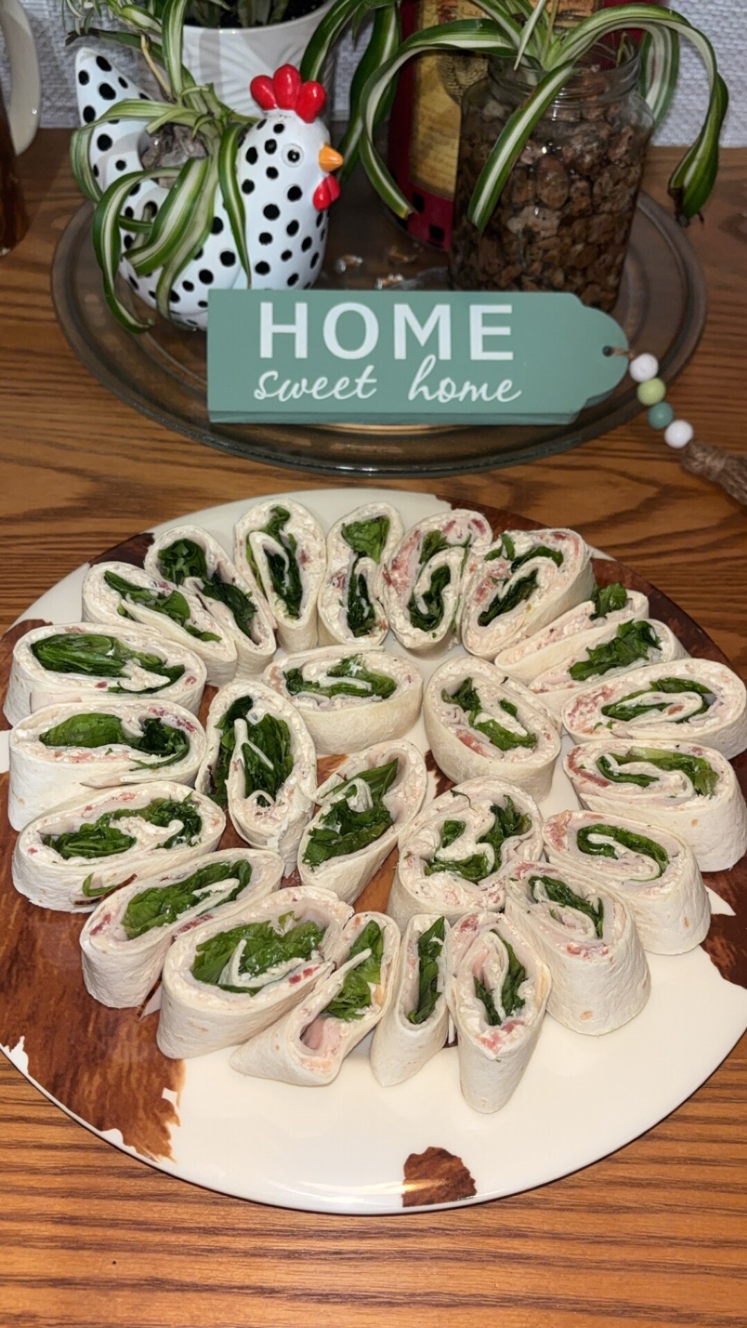 A plate of tortilla pinwheels filled with spinach and deli meat is arranged in a circle on a brown and white plate. A Home sweet home sign and a plant are in the background.