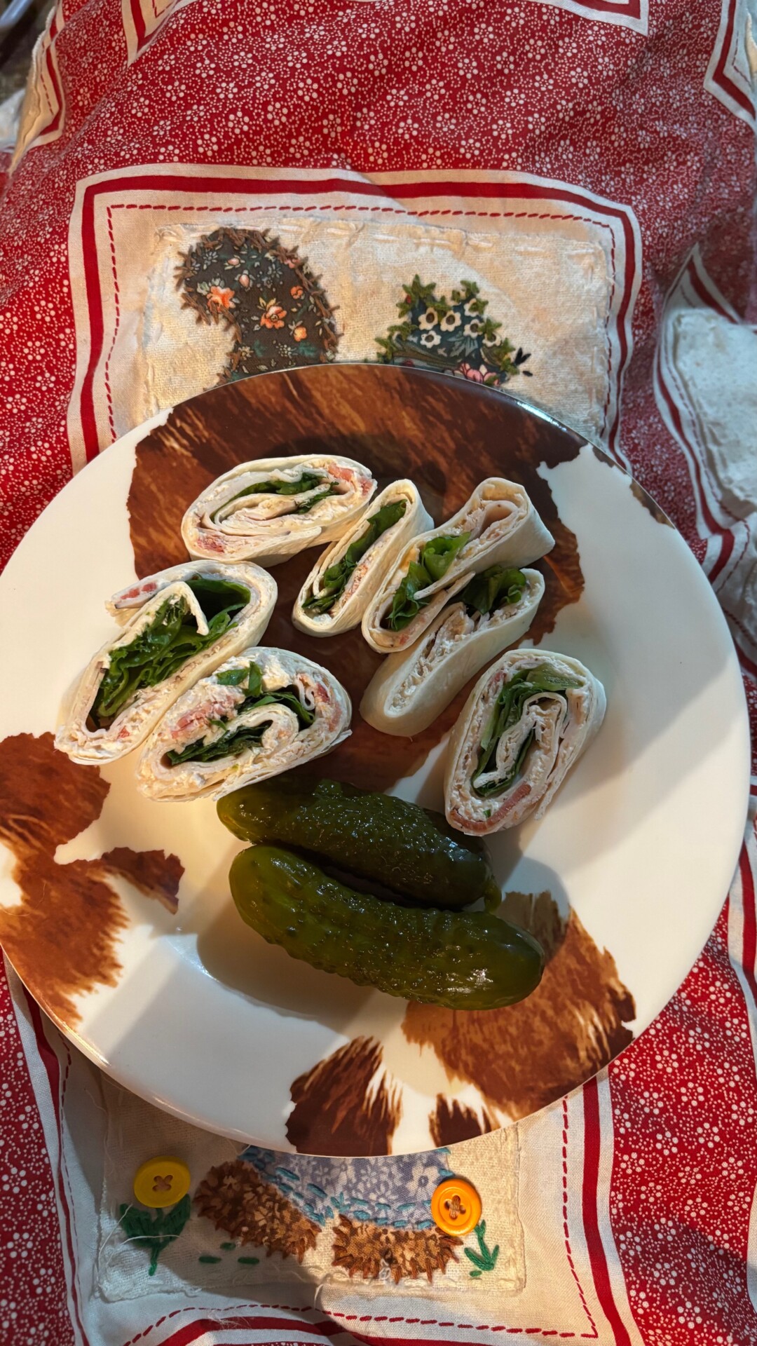 A plate with six tortilla pinwheel sandwiches filled with greens and deli meat, and two pickles, placed on a red and white patterned cloth with a festive design.