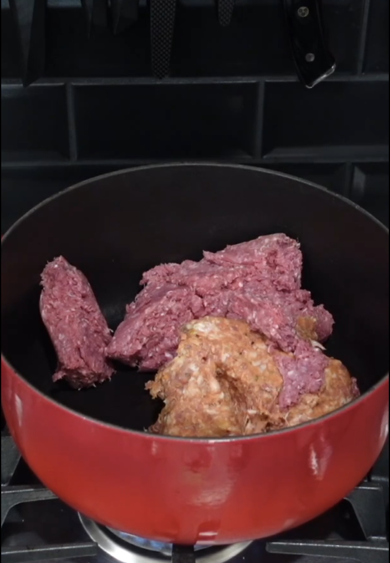 Raw ground meat in a red pot on a stovetop, ready to be cooked. Black tile backsplash and hanging knives are visible in the background.