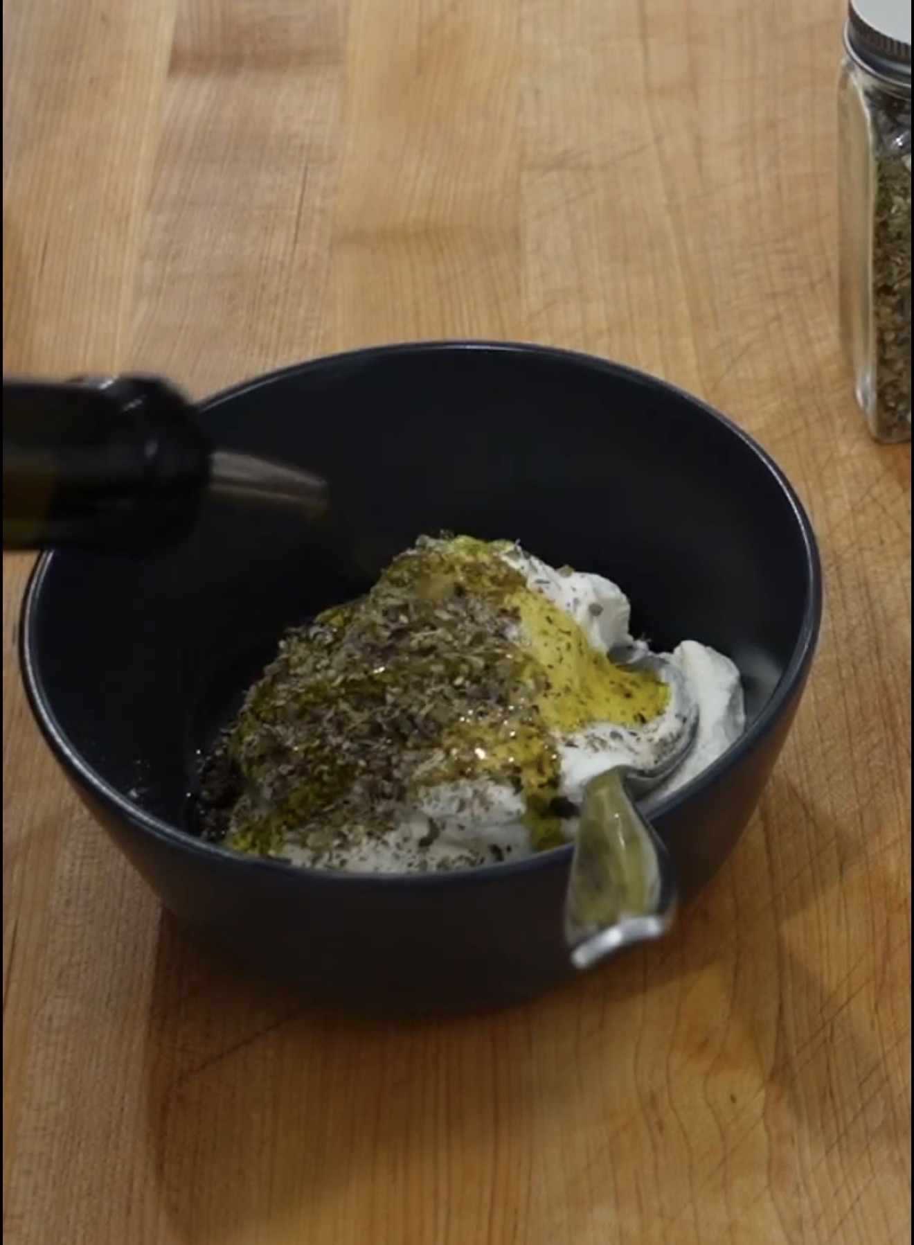 A dark bowl with yogurt, olive oil, and herbs sits on a wooden surface. A bottle pours more olive oil onto the mixture. A jar of dried herbs is visible nearby.
