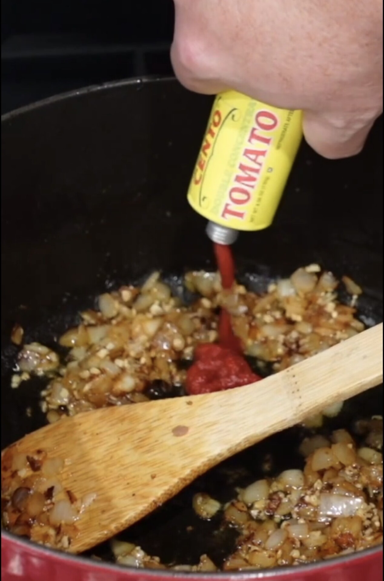 A hand squeezes tomato paste from a yellow tube into a pot with sautéed onions and garlic. A wooden spoon rests inside the pot.