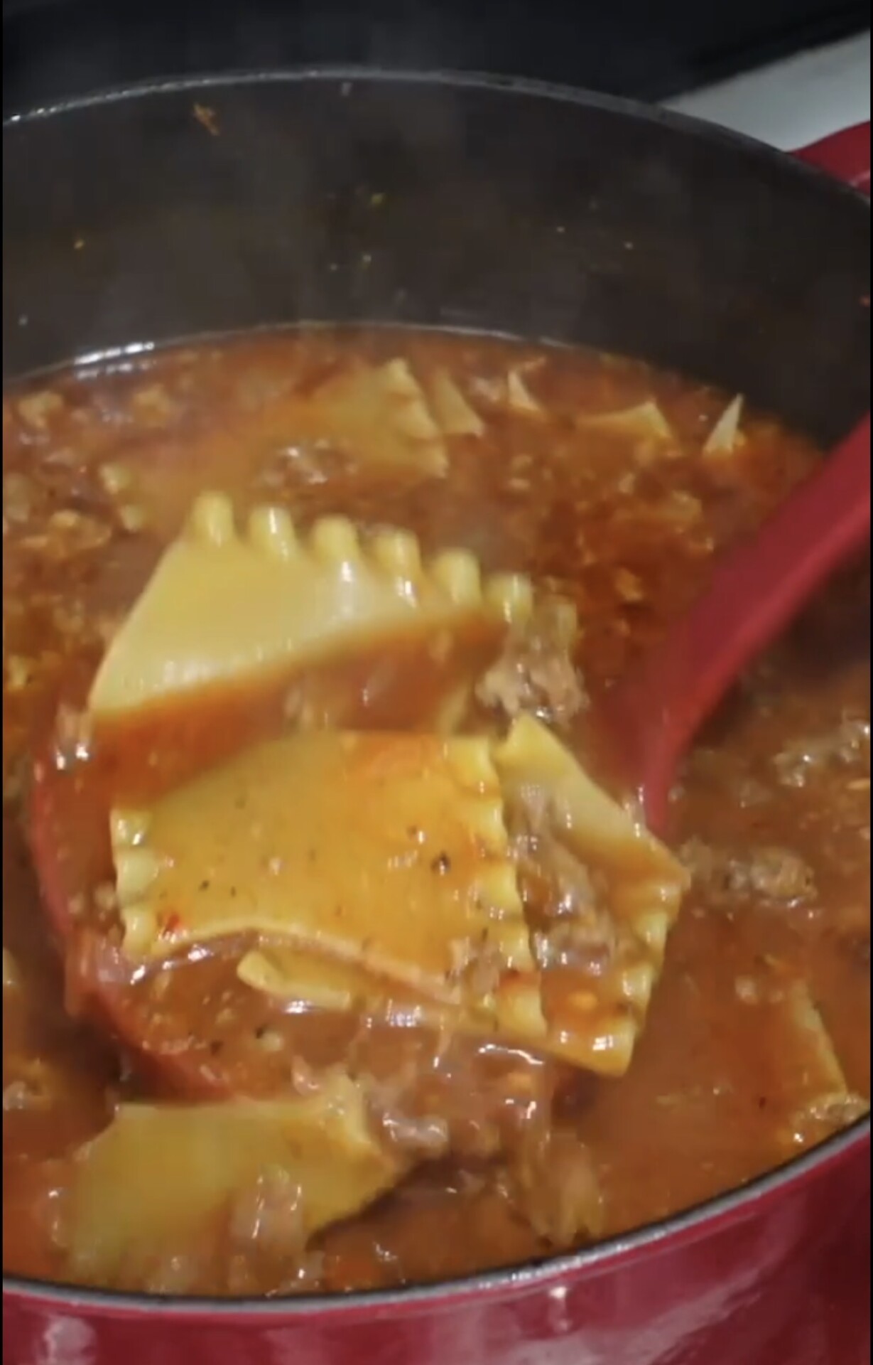 A red pot filled with lasagna soup, showing broken lasagna noodles, ground meat, and tomato-based broth. A red ladle is lifting a portion of the soup. Steam rises from the pot.
