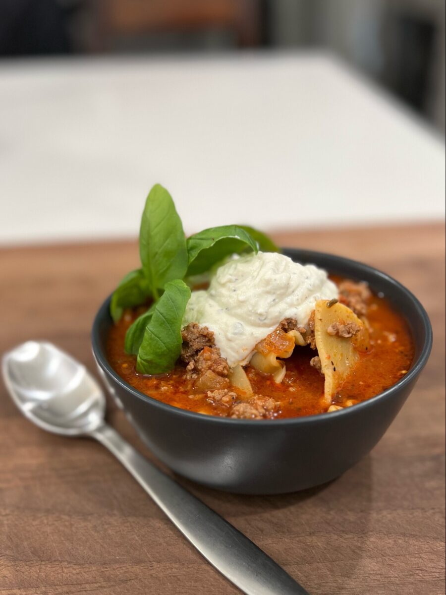 A black bowl of hearty soup with ground meat, pasta, fresh basil, and a large dollop of creamy white topping, placed on a wooden surface next to a metal spoon.