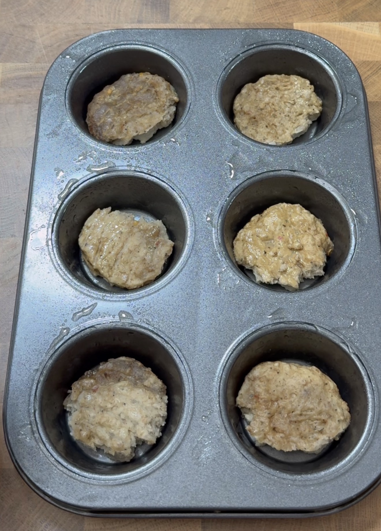 A muffin tin with six cups, each holding a cooked, round patty that looks like sausage or meatloaf, on a wooden surface.