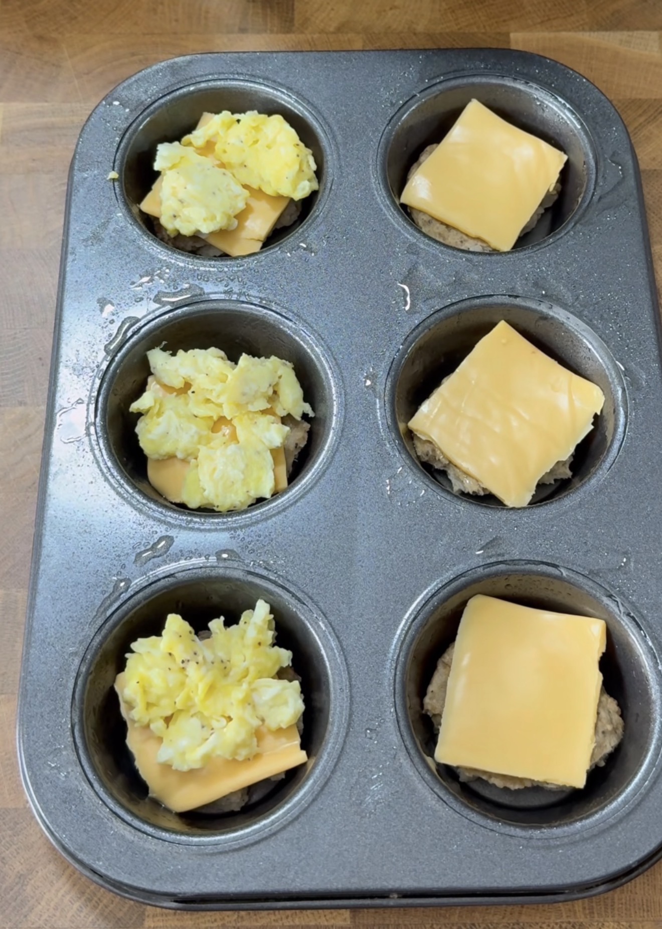 A muffin tin with six wells, each containing a breakfast sandwich layer: sausage patty, scrambled eggs, and a slice of melted cheese on top, on a wooden surface.