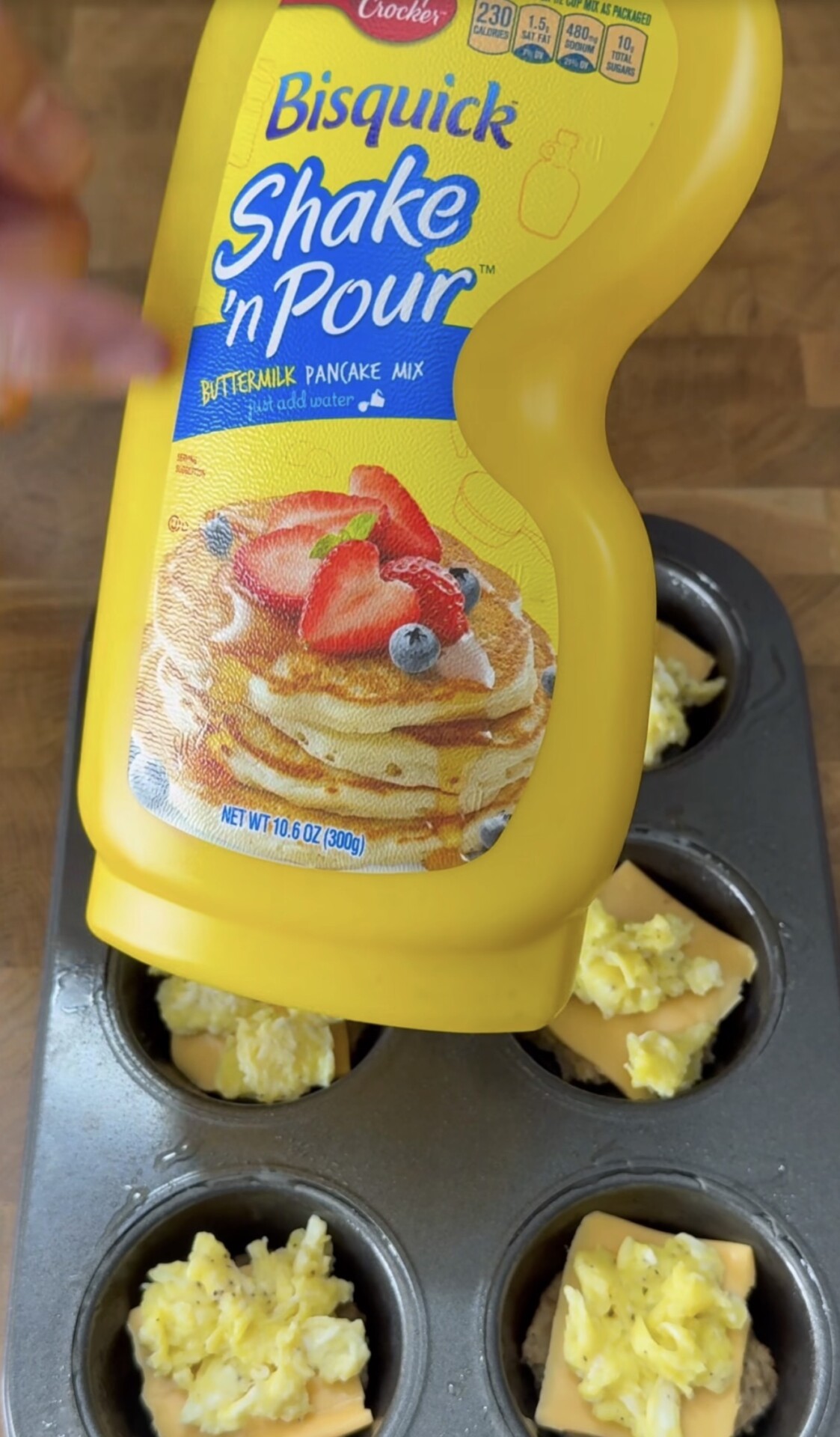 A hand pours Bisquick Shake n Pour pancake mix over muffin tin cups filled with scrambled eggs and cheese slices, sitting on a wooden surface.