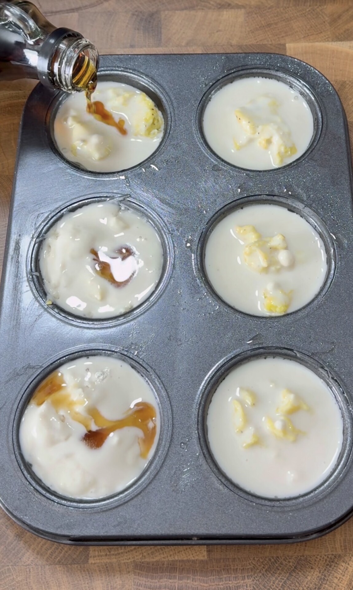 A muffin tin with six cups filled with batter and some chunks. Liquid is being poured into one of the cups from a bottle, on a wooden surface.