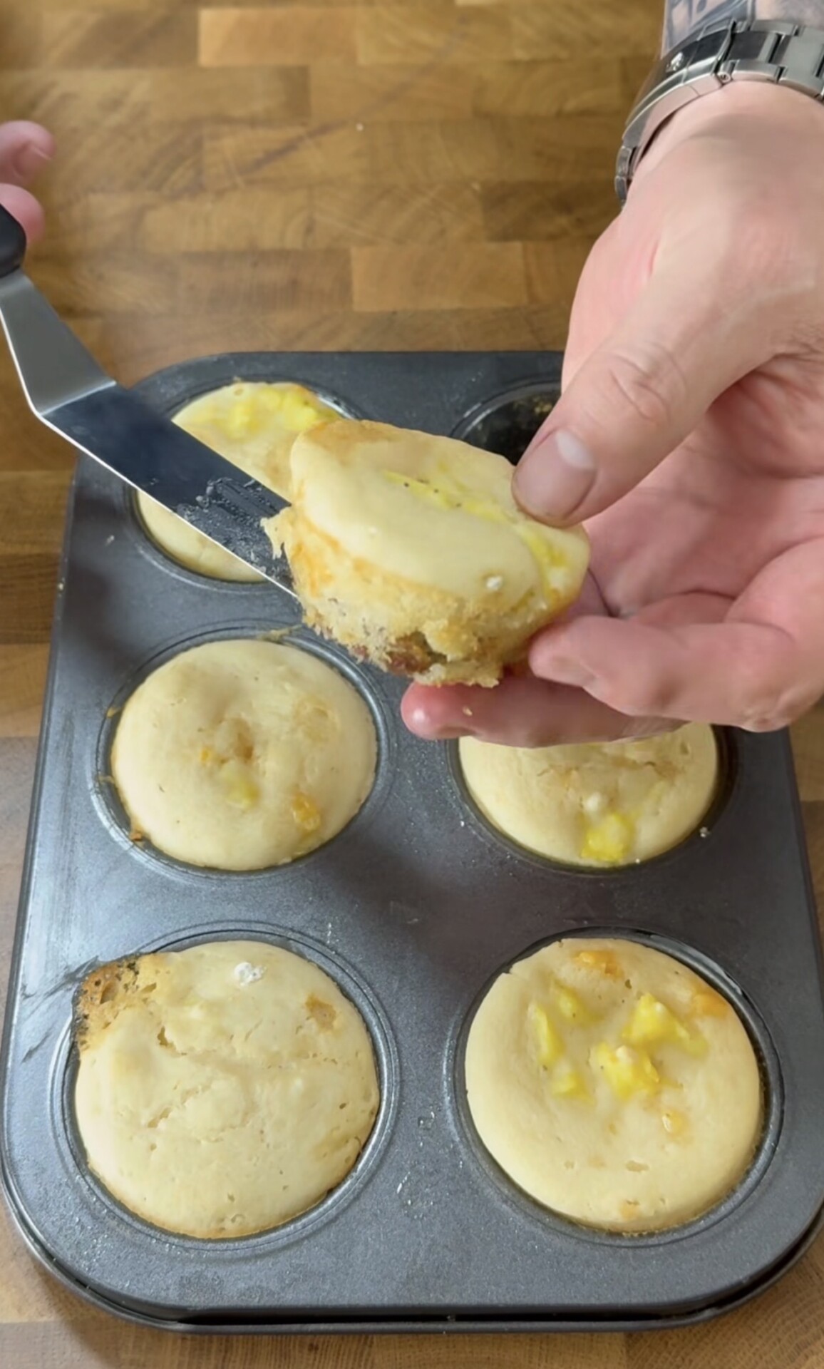 A hand lifts a muffin from a metal muffin tin using a spatula. The muffins are light in color with small yellow pieces inside. The background is a wooden surface.