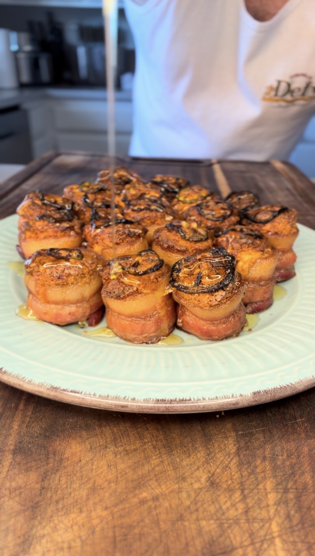 A person drizzles sauce over a plate of bacon-wrapped appetizers, neatly arranged on a light green plate atop a wooden table in a kitchen setting.