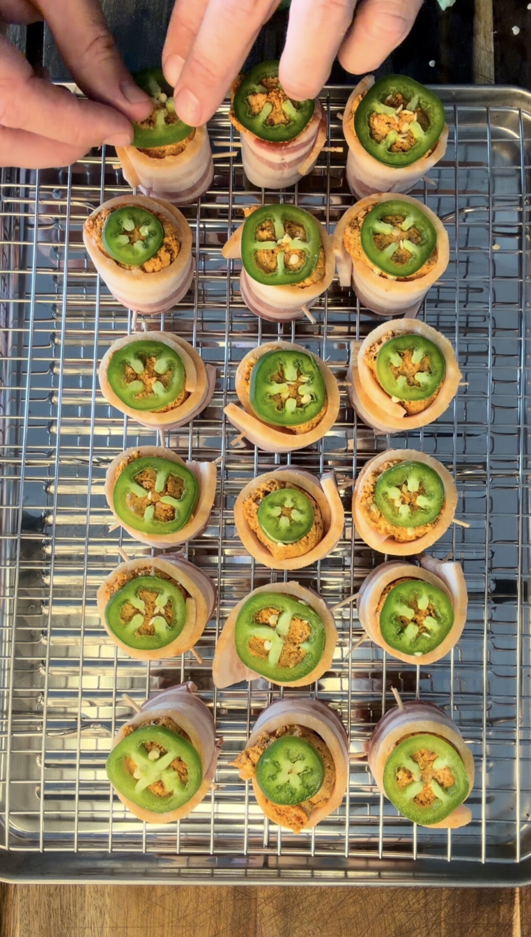 Hands arrange bacon-wrapped appetizers topped with jalapeño slices on a wire rack over a baking sheet. Each piece is neatly aligned in rows, showing the green jalapeño rings and light brown bacon spirals.