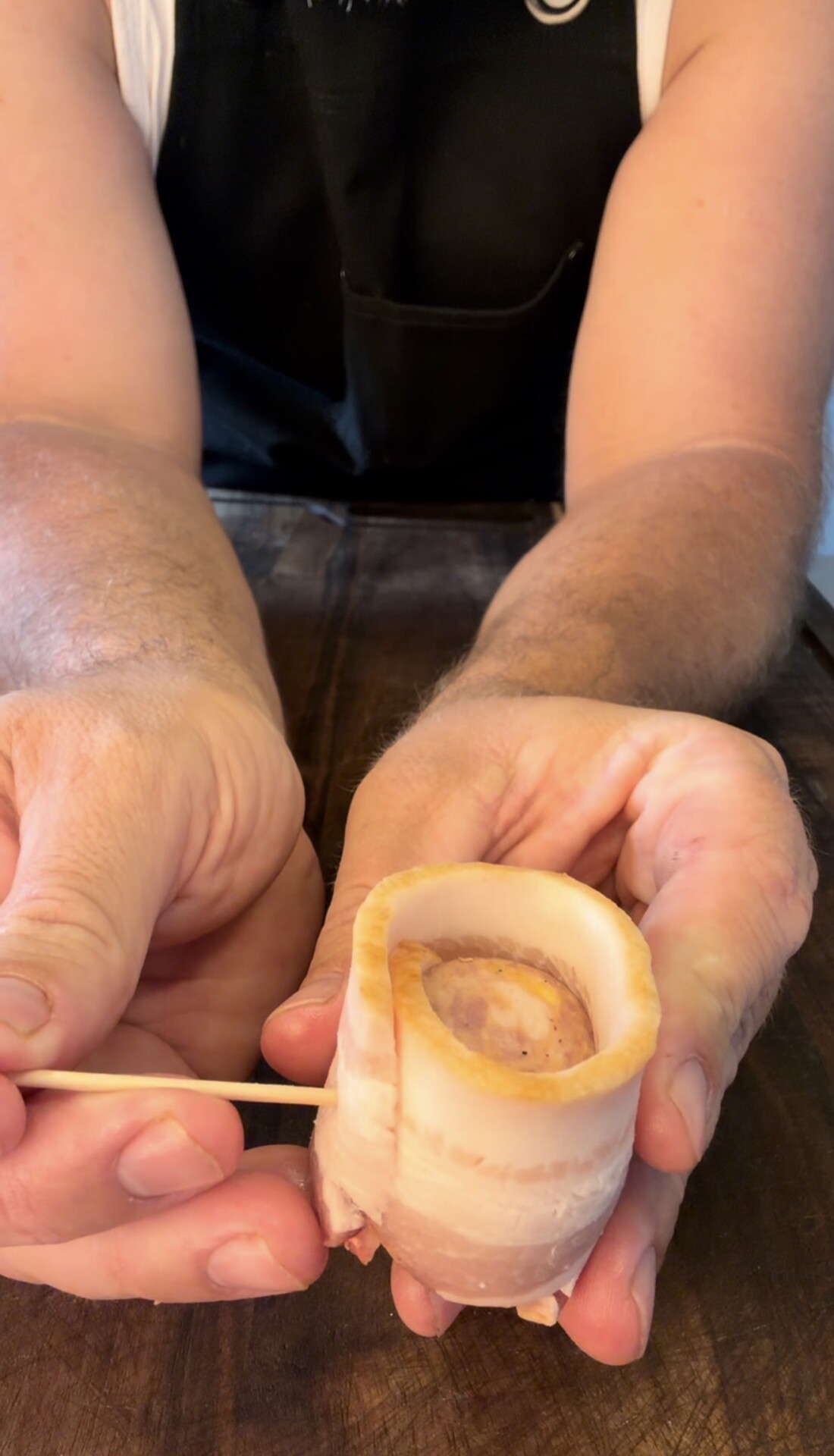 A person holding a piece of marrow bone wrapped in bacon, using a wooden skewer to secure it, on a wooden table.