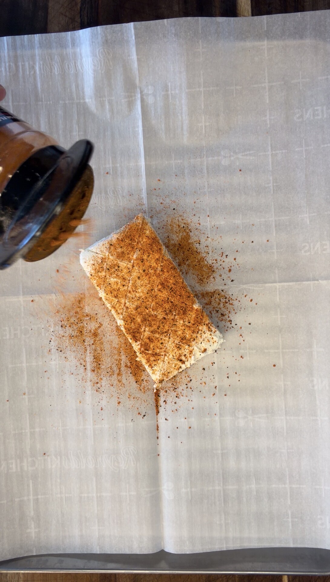A hand sprinkles reddish-brown seasoning onto a rectangular block of tofu placed on a sheet of parchment paper, with spice scattered around the tofu.