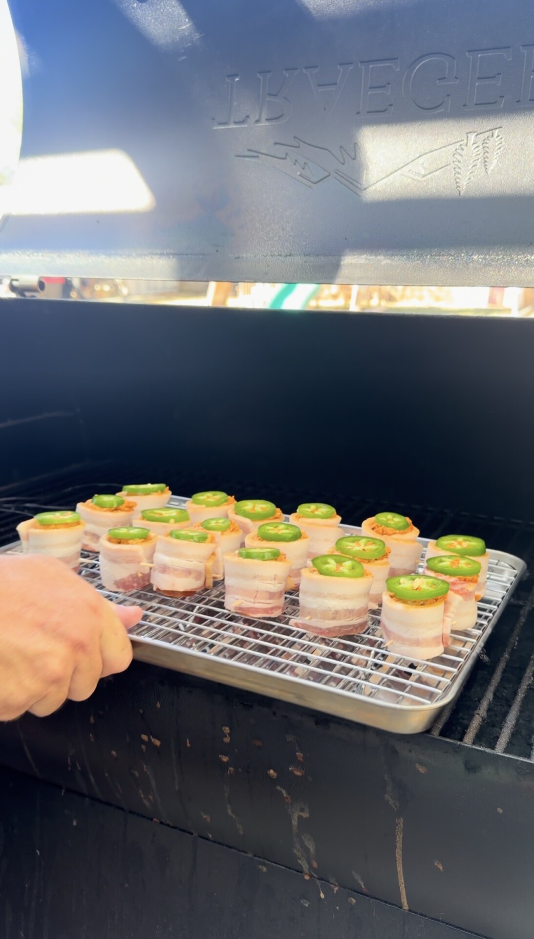 A hand places a rack of bacon-wrapped appetizers topped with jalapeño slices into a smoker grill.