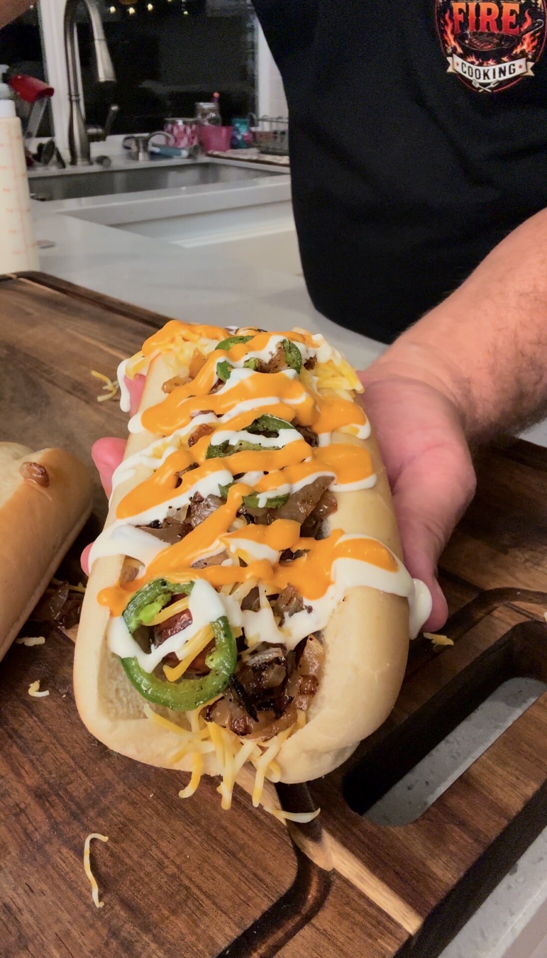 A hand holds a loaded sandwich topped with sliced jalapeños, grilled meat, shredded cheese, and drizzles of white and orange sauce on a wooden cutting board.