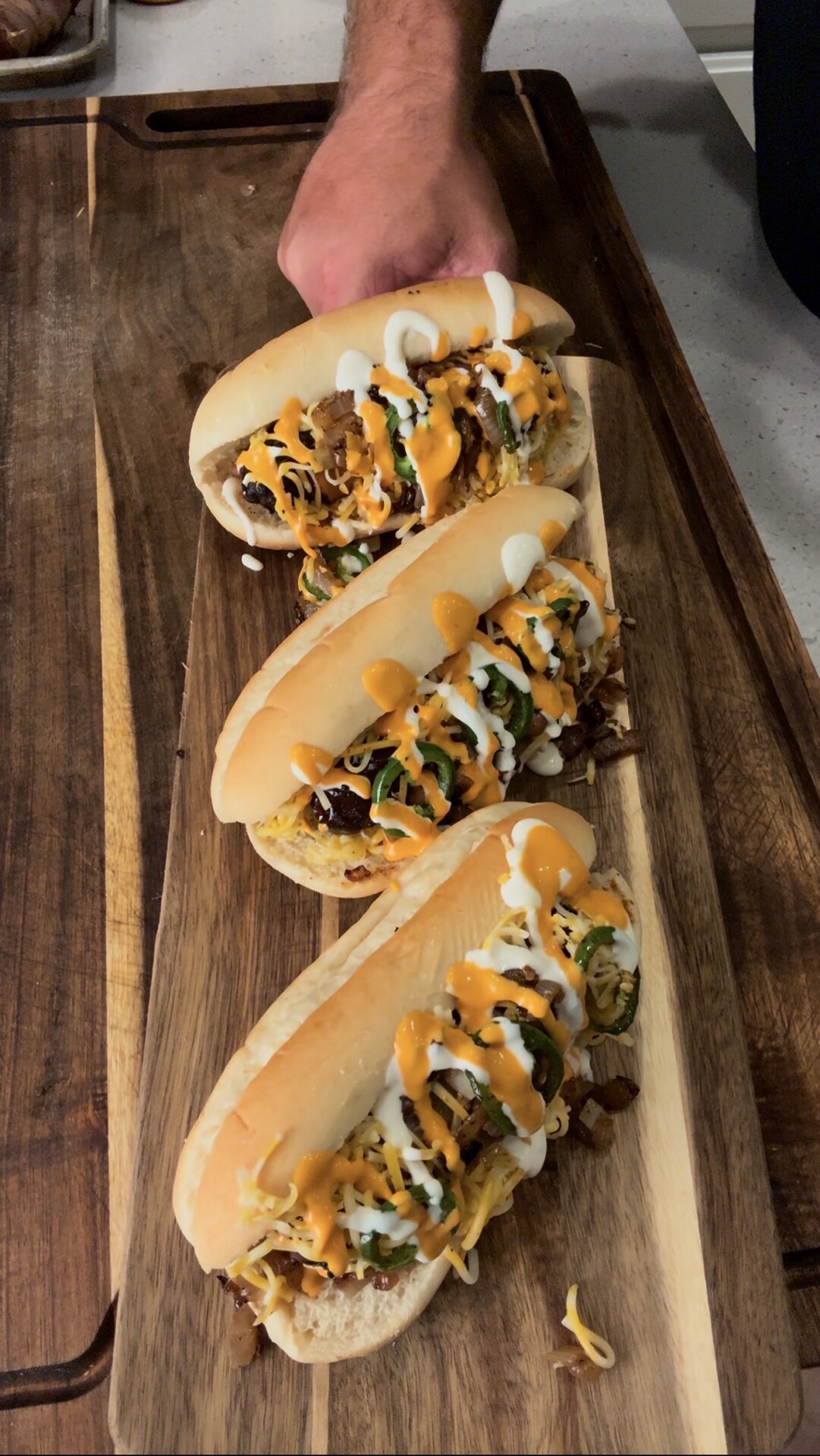 Three loaded hot dogs on a wooden board, topped with cheese, jalapeños, diced onions, and drizzled with orange and white sauces. A hand holds the hot dog at the top of the image.