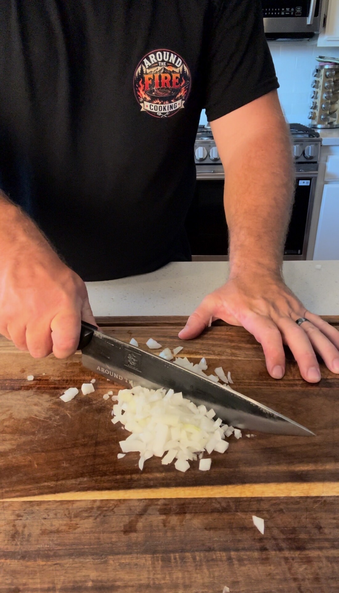A person chops a white onion on a wooden cutting board in a kitchen, holding a large knife and wearing a black T-shirt with a colorful logo.