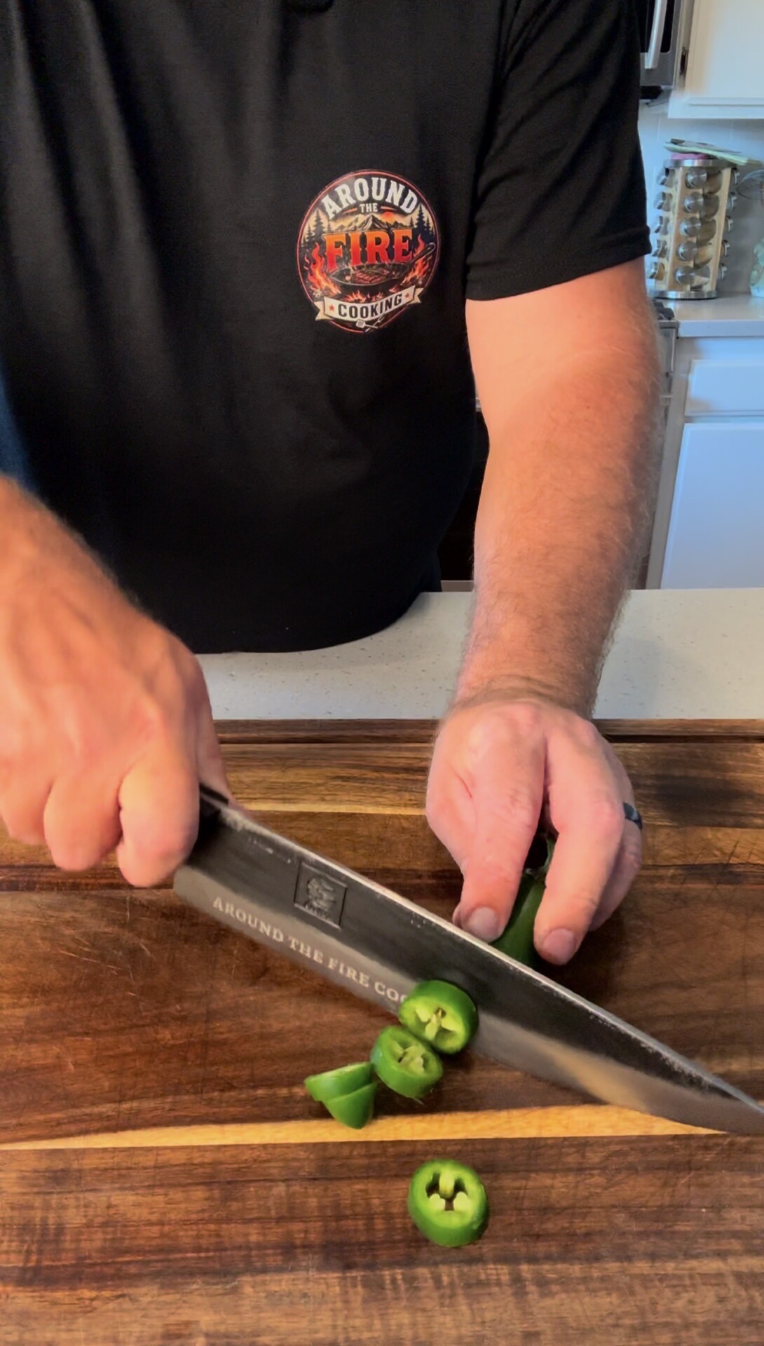 A person slices green jalapeños on a wooden cutting board with a large knife. The person wears a black shirt with a colorful Around the Fire Cooking logo. A kitchen counter and white cabinets are in the background.