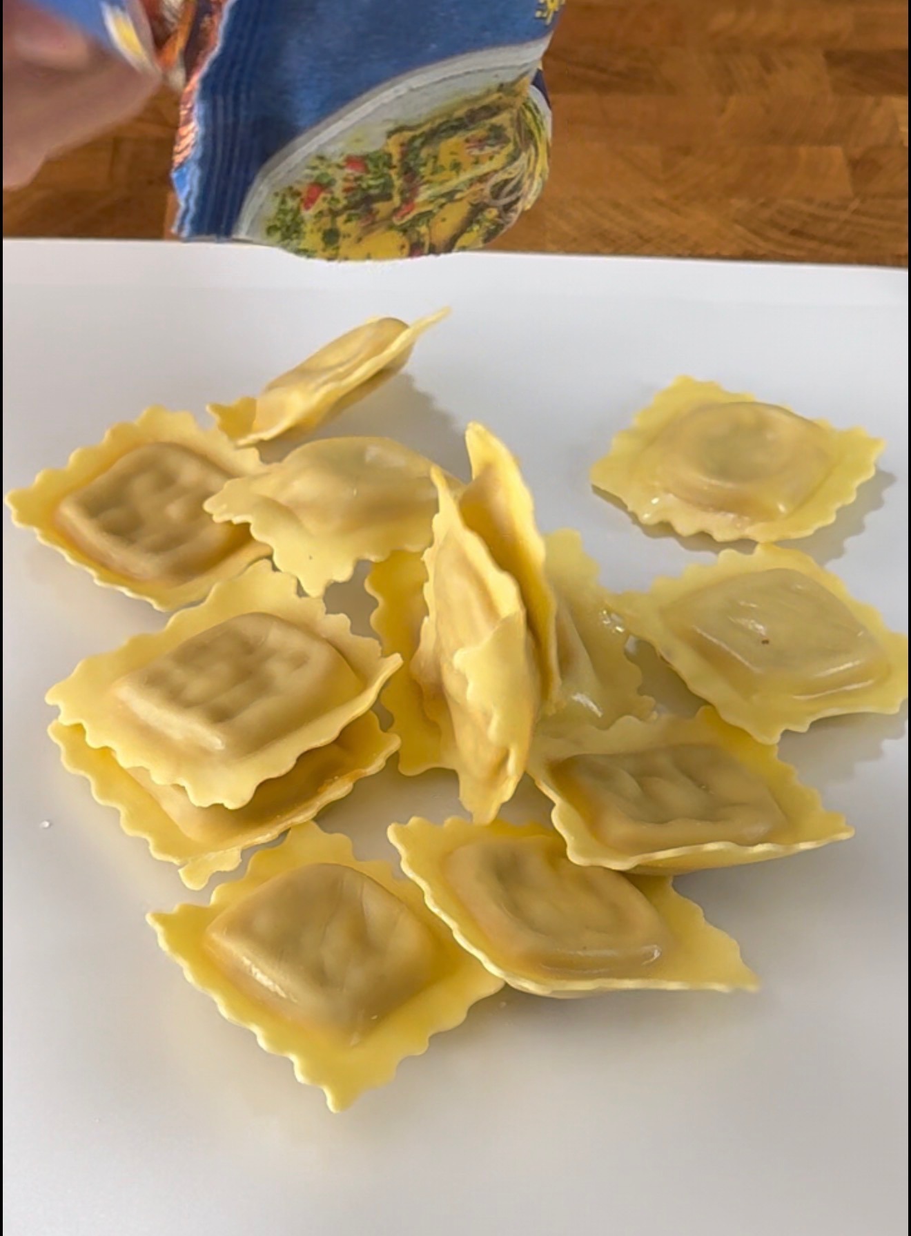 Uncooked square ravioli pieces being poured from a package onto a white surface, with a hand holding the package partially visible in the upper left corner.