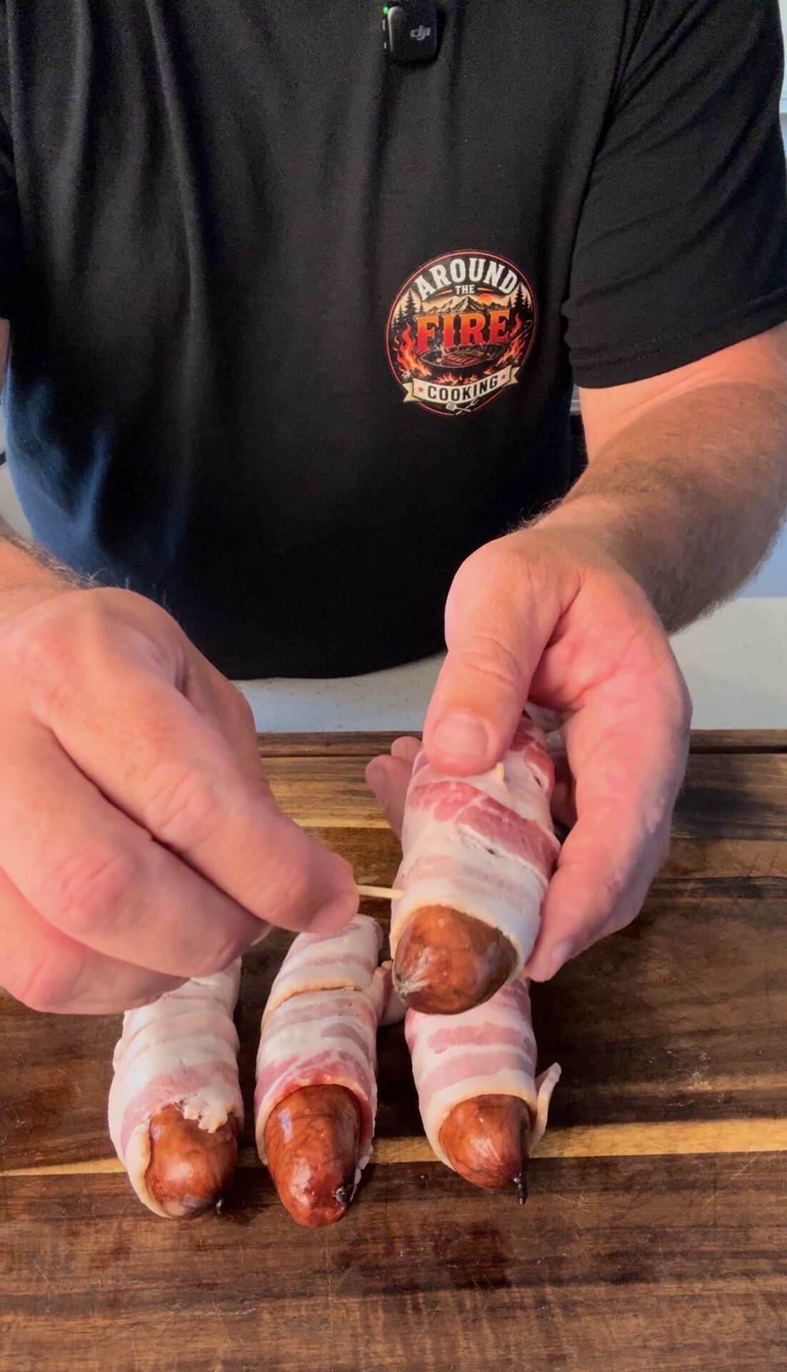 A person wraps bacon around sausages and secures them with toothpicks on a wooden surface. The person is wearing a black Around the Fire barbecue t-shirt.