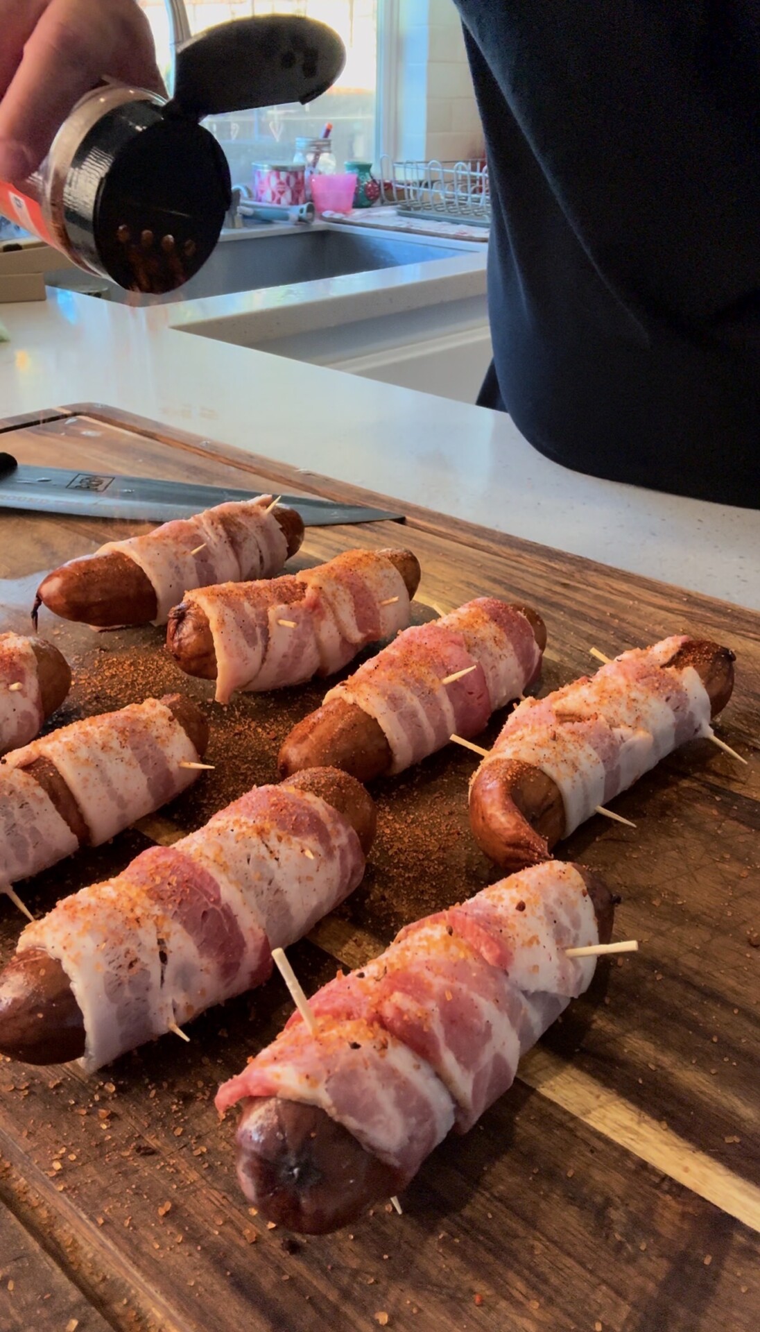 Several sausages wrapped in bacon are arranged on a wooden board, secured with toothpicks, as seasoning is sprinkled over them. A kitchen counter and sink are visible in the background.
