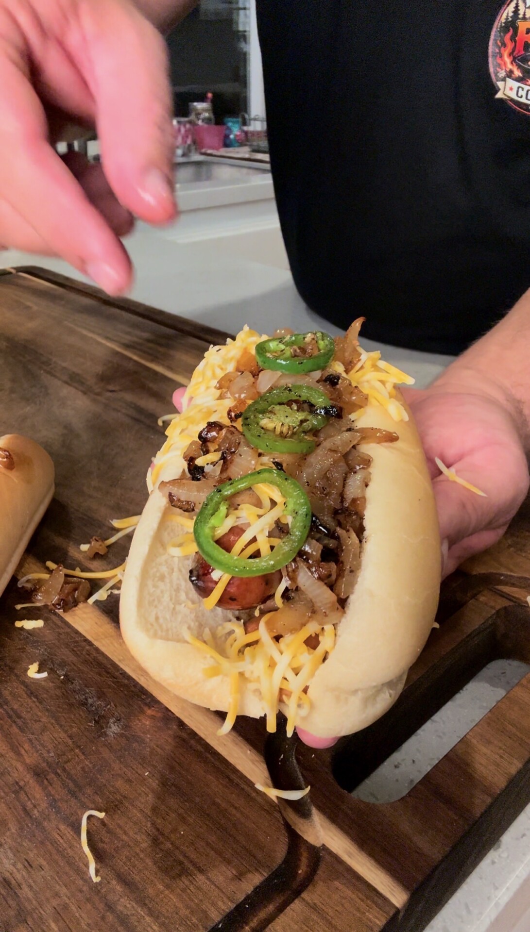 A person holds a hot dog topped with shredded cheese, grilled onions, and sliced jalapeños on a wooden surface.