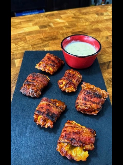 Six grilled, bacon-wrapped cheese bites are arranged on a dark slate serving board, with a small red bowl of creamy dipping sauce placed at the top right corner.