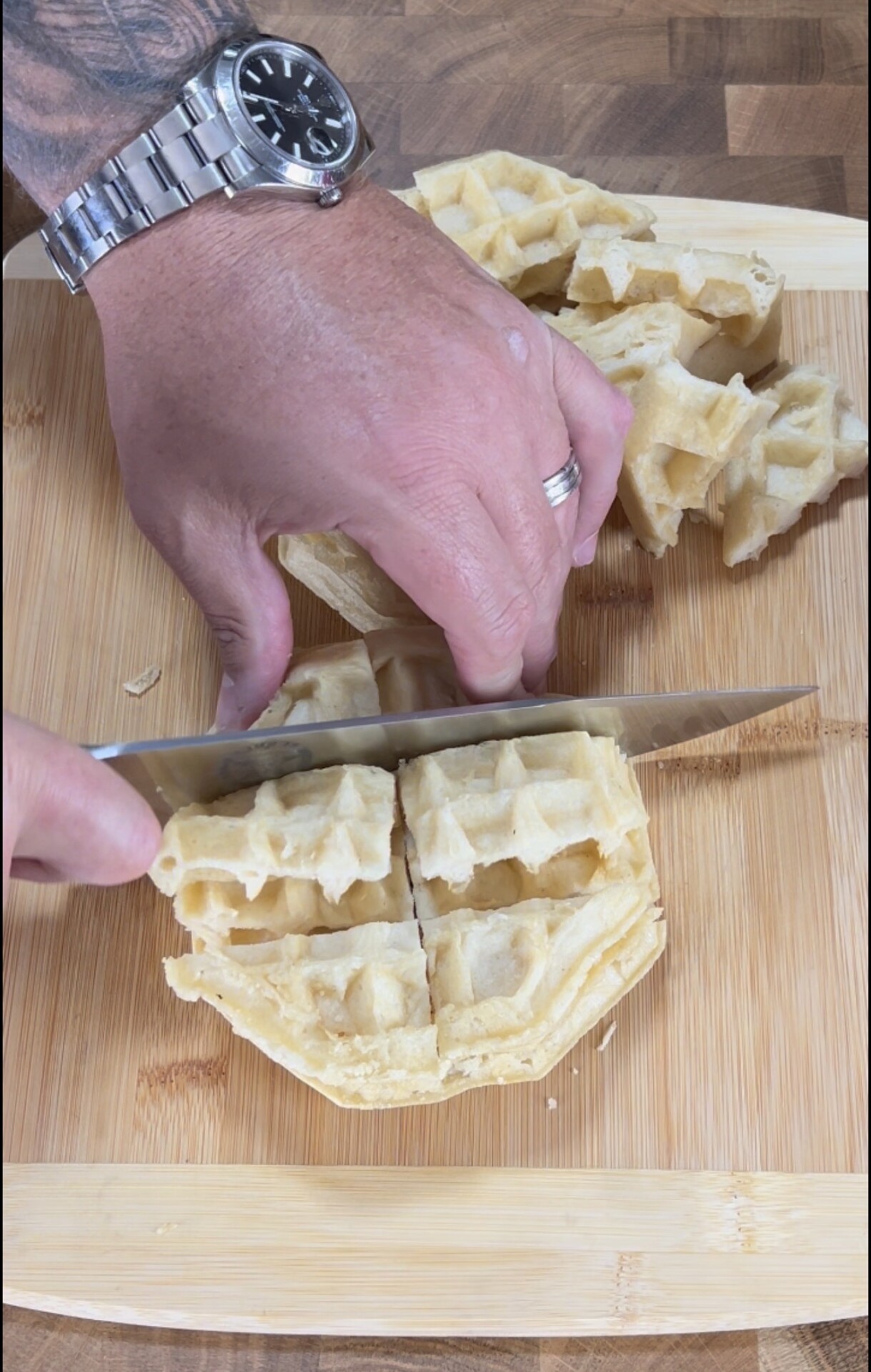 A person wearing a watch and ring slices a waffle into quarters on a wooden cutting board using a large knife.