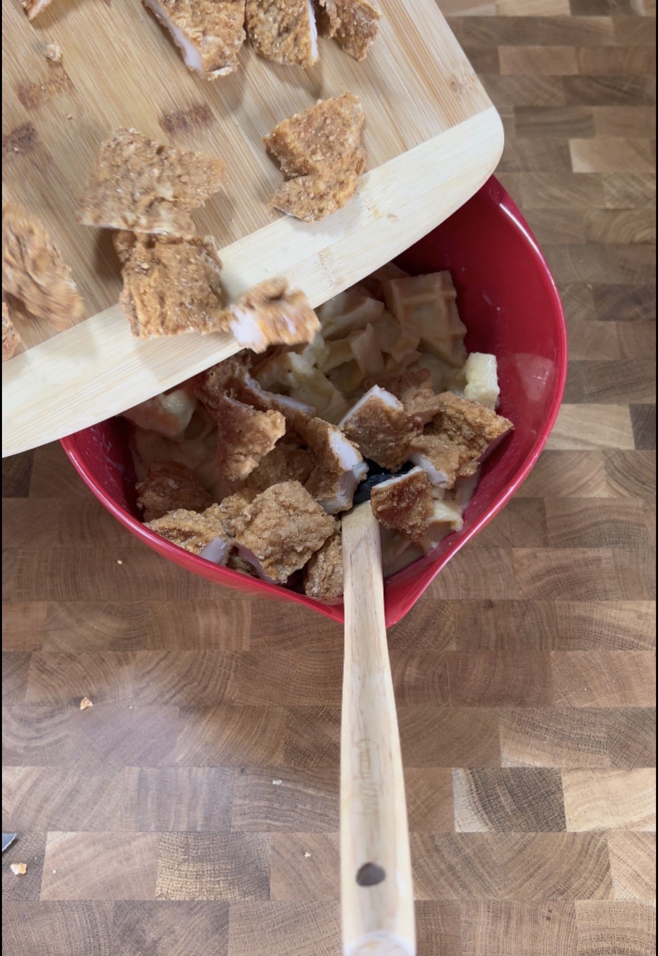 Chunks of breaded chicken are being poured from a wooden cutting board into a red mixing bowl filled with chopped apples. A wooden spoon rests in the bowl on a wooden countertop.