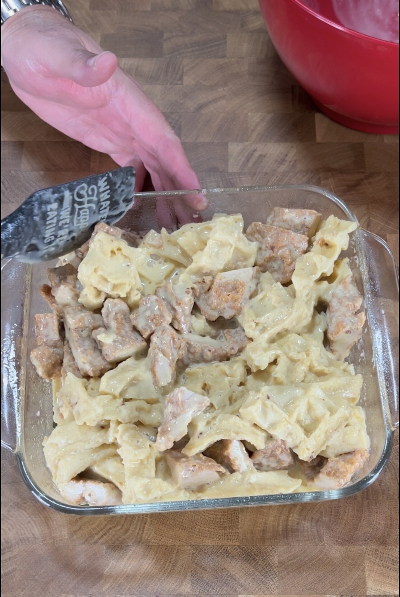 A hand holds a spatula over a glass dish filled with bowtie pasta and pieces of sausage in a creamy sauce on a wooden surface.