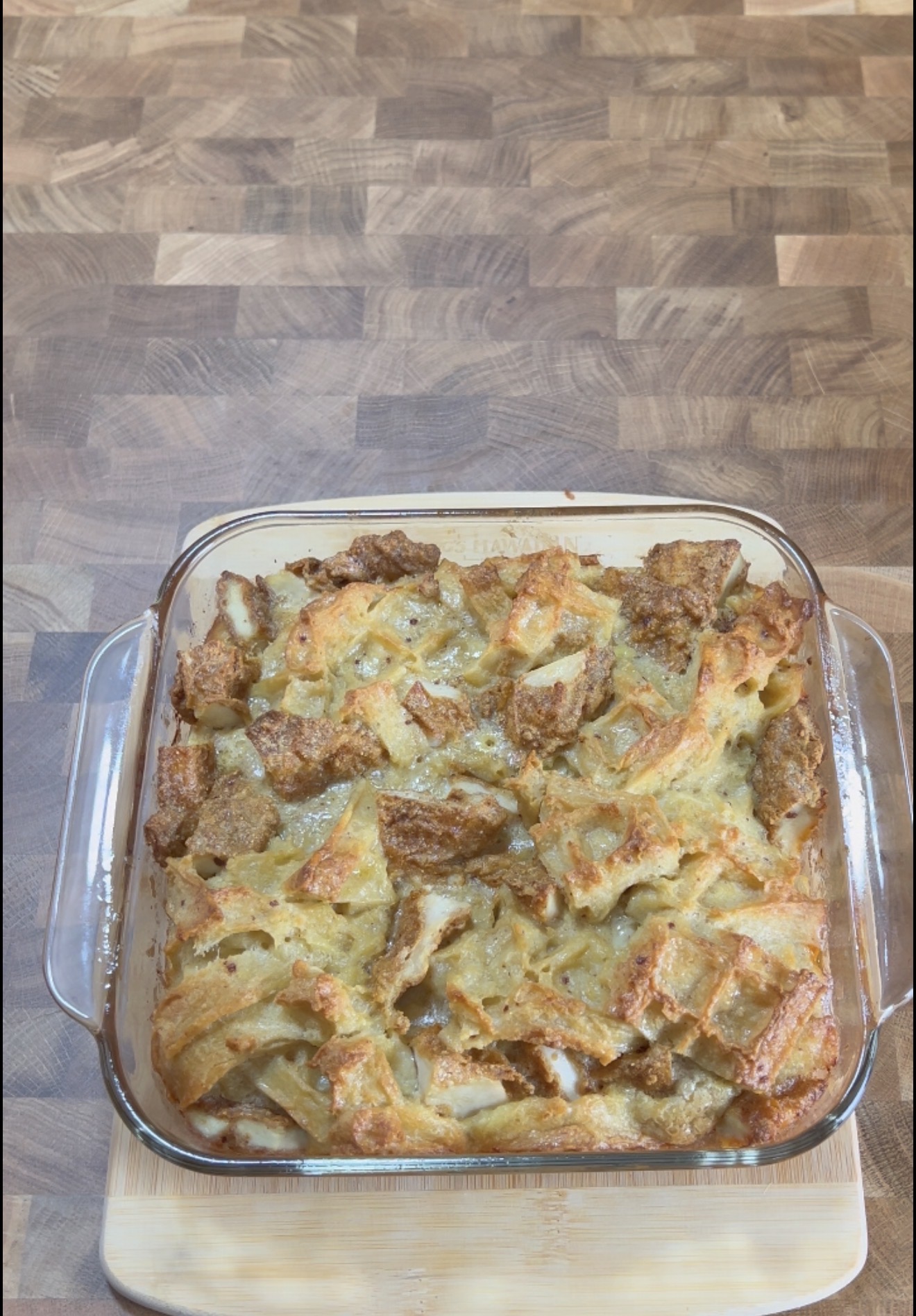 A glass baking dish filled with golden-brown bread pudding sits on a light wooden cutting board atop a patterned wooden countertop.