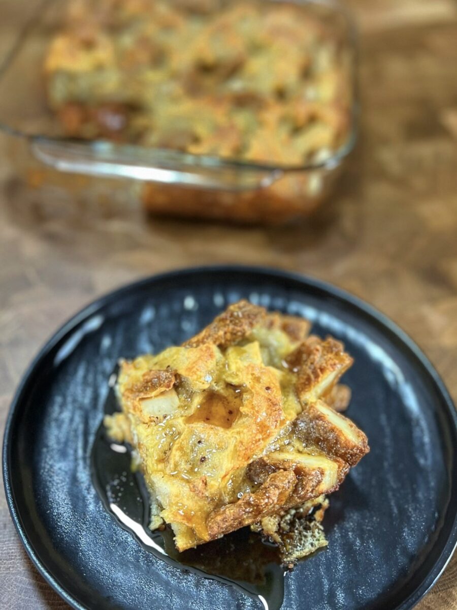 A slice of bread pudding drizzled with syrup sits on a black plate, with a glass baking dish containing more bread pudding in the background.