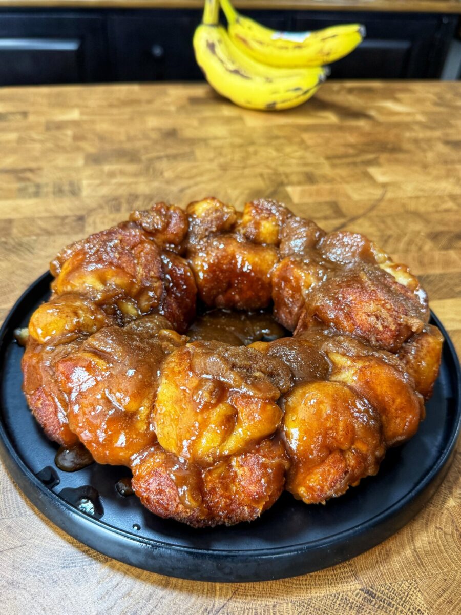 A round monkey bread covered in caramel sauce sits on a black plate on a wooden countertop, with a bunch of bananas in the background.