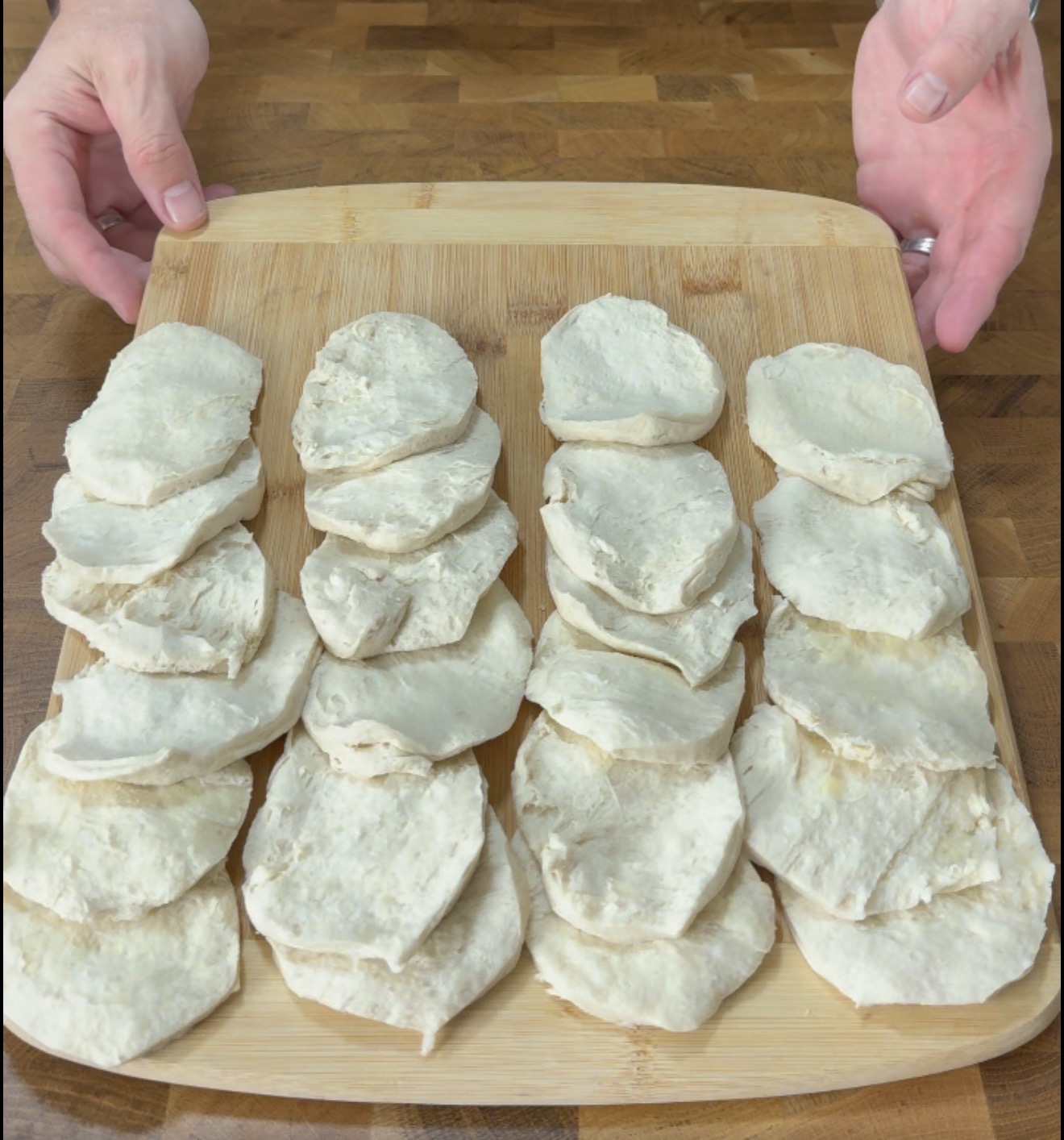 Four neat rows of uncooked dough pieces are arranged on a wooden cutting board, with two hands visible on either side of the board, set on a wooden surface.