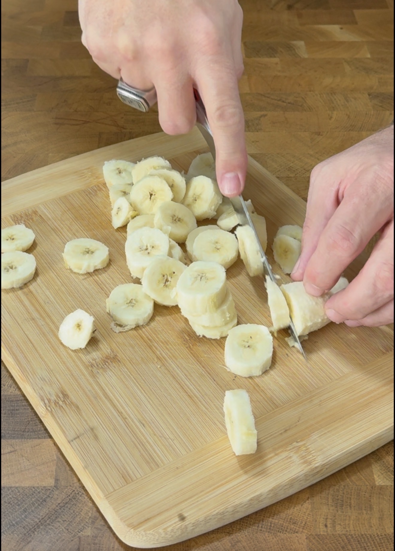 A person slices a banana on a wooden cutting board with a knife. Banana slices are scattered on the board, and the person wears a ring on one finger.