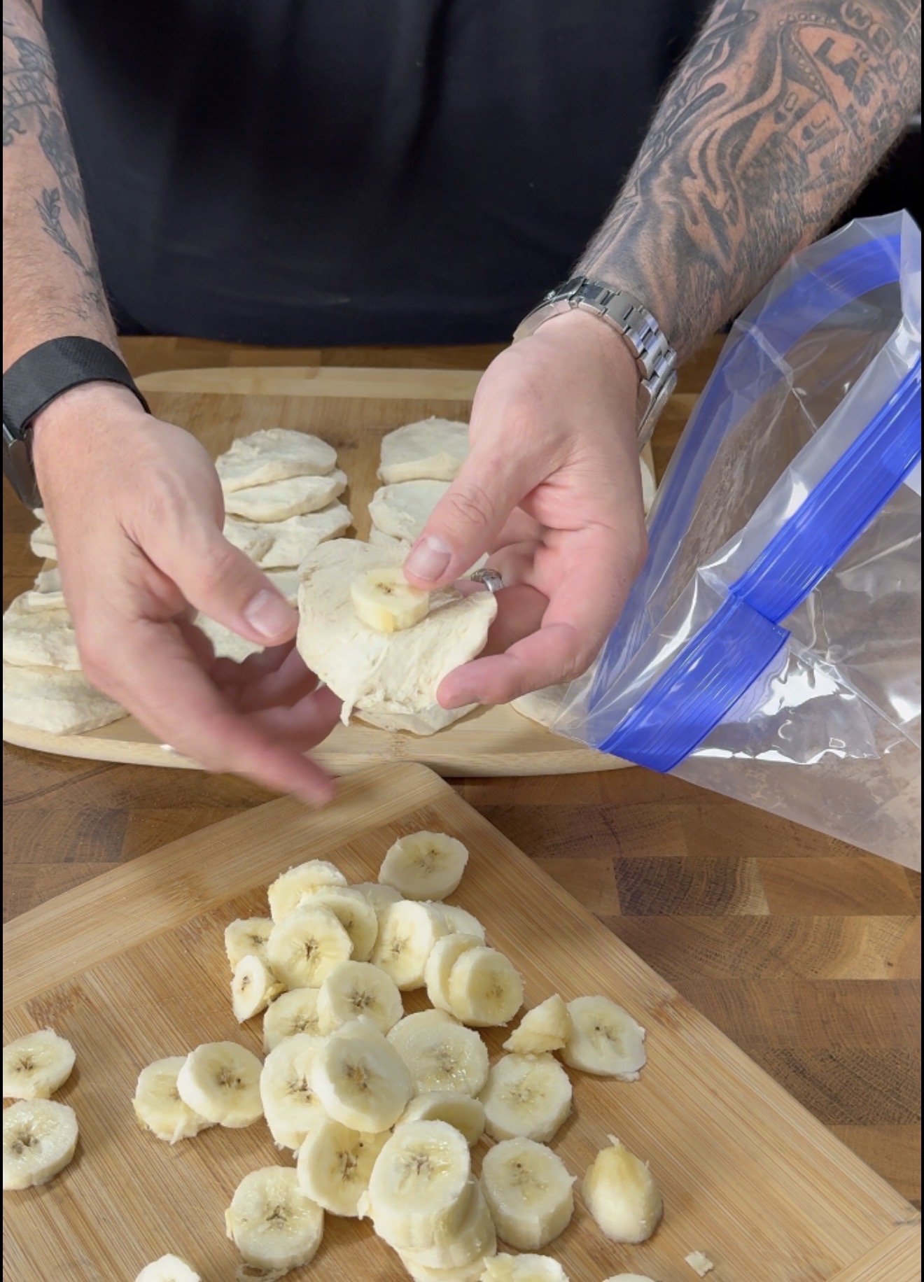 Tattooed hands place banana slices between biscuit dough rounds on a wooden board, with more banana slices and a plastic bag nearby.