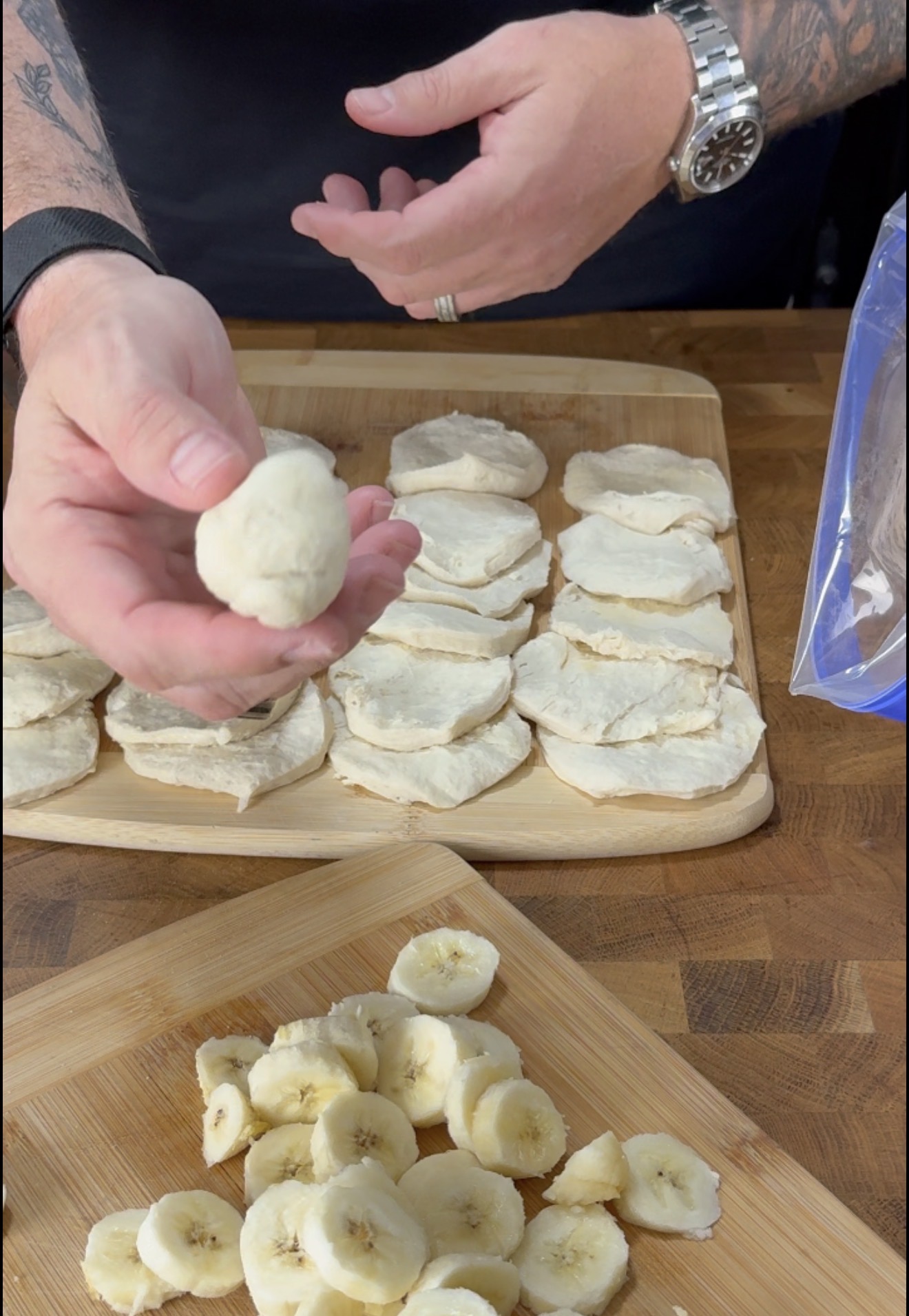 A person holds a dough ball over sliced biscuits, with banana slices on a wooden board and more biscuit slices on a cutting board nearby.