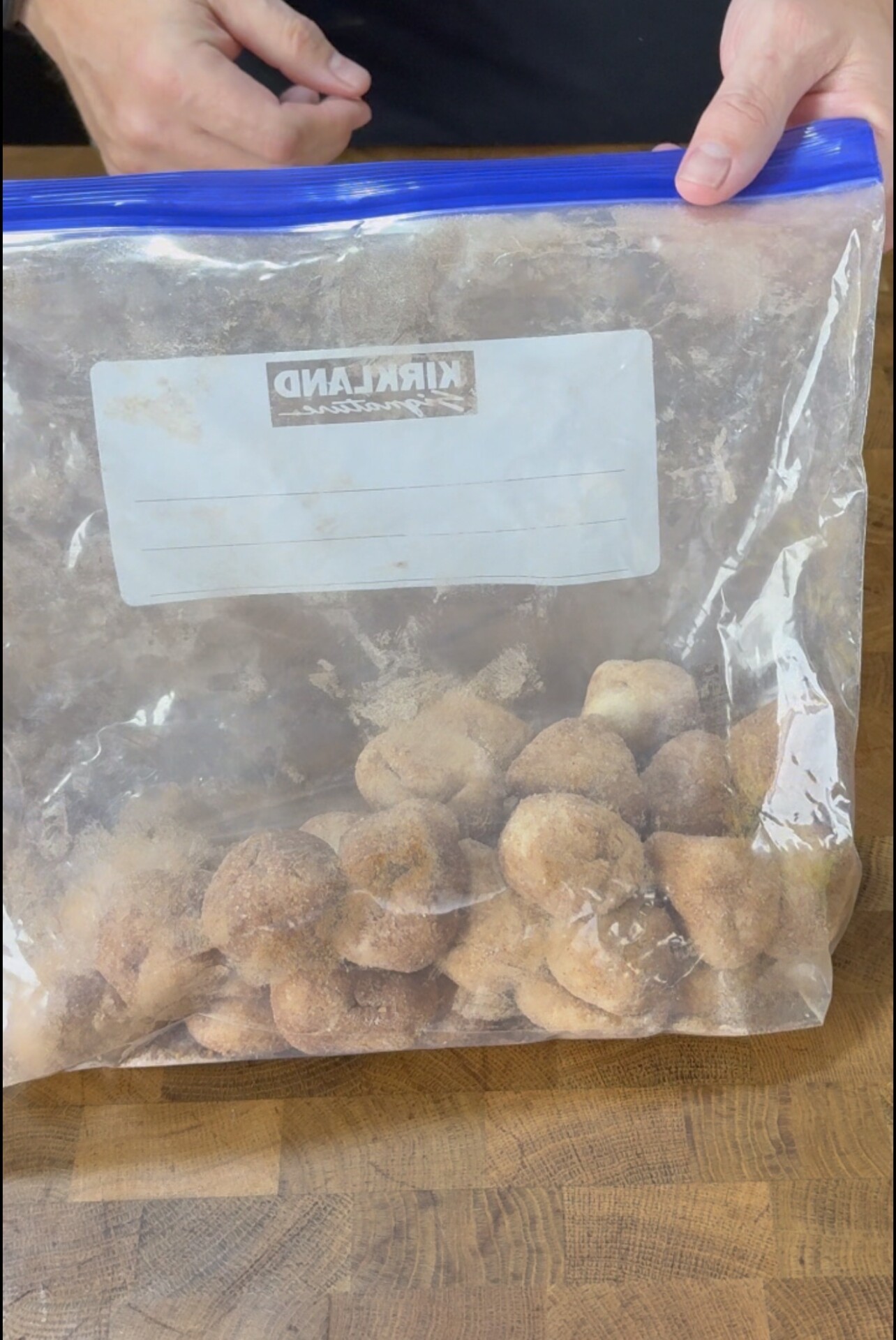 A person holds a large zipper bag labeled Kirkland filled with small, round, cinnamon-sugar coated dough balls on a wooden surface.