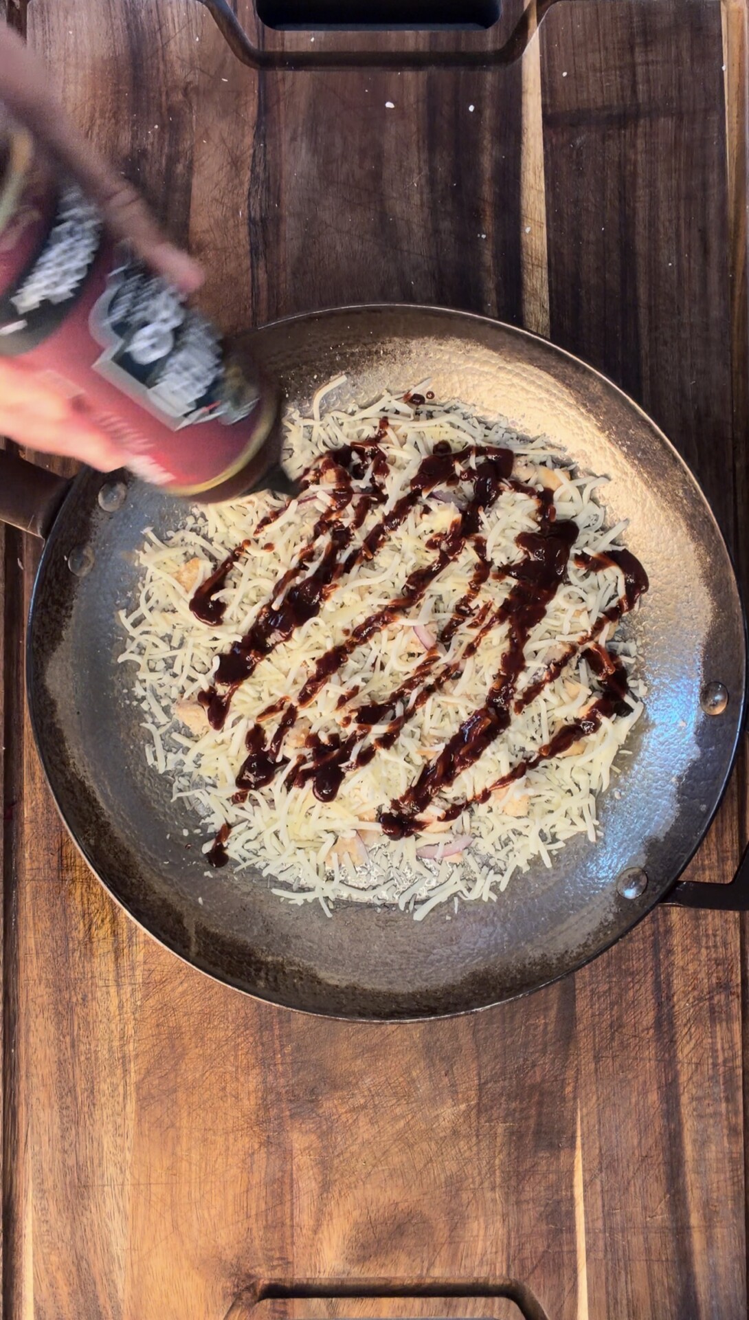 A hand pours barbecue sauce in zigzag lines over shredded cheese on a tortilla in a pan on a wooden surface.