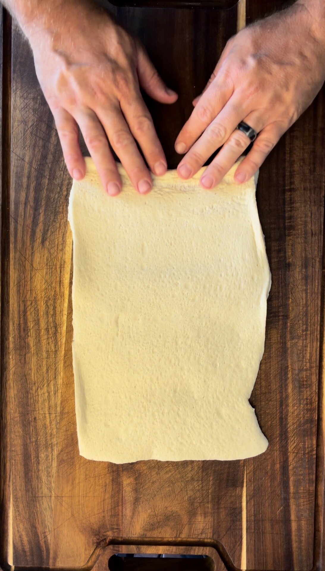 A person with a ring on their left hand rolls out rectangular dough on a wooden surface.