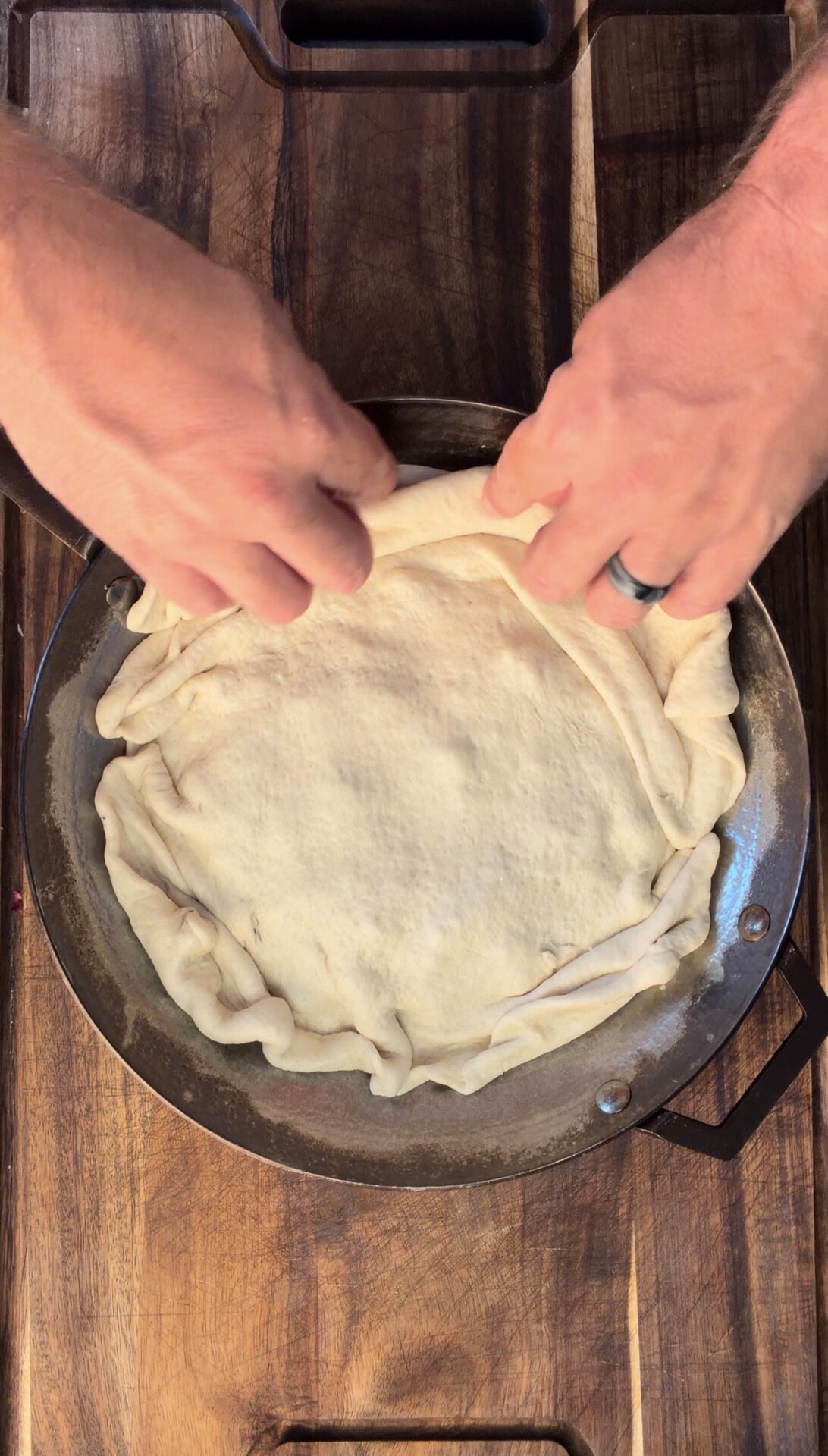 Two hands folding the edge of raw dough inside a round pan on a wooden surface, preparing it for baking or filling.