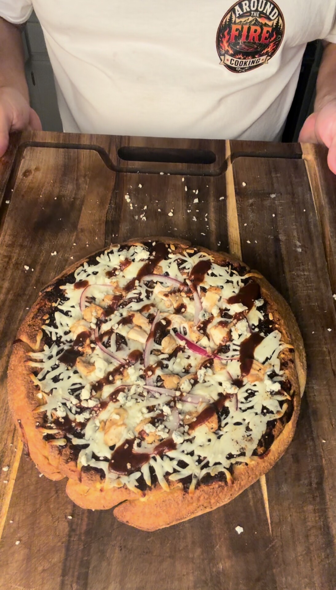 A person in a white shirt holds a wooden board with a homemade pizza topped with cheese, onions, and dark sauce.