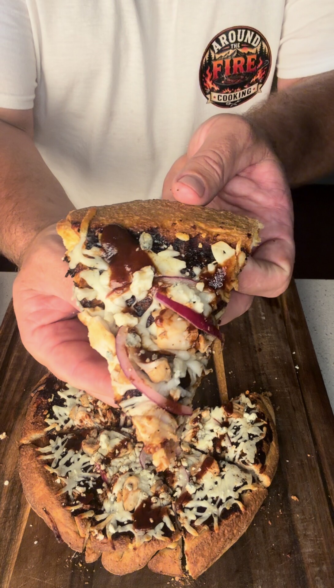 A person holds a slice of pizza with barbecue sauce, red onion, cheese, and chicken, above a wooden cutting board with the remaining pizza. The person wears a white shirt with an Around the Fire Cooking logo.