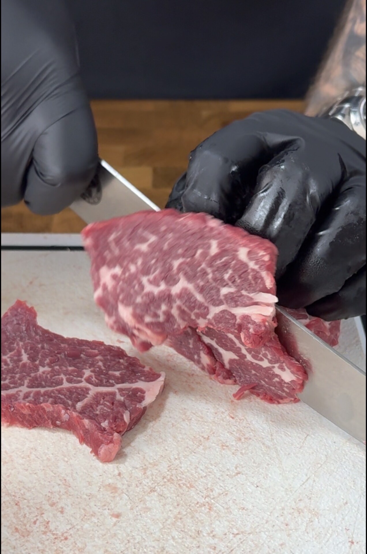 A person wearing black gloves slices a piece of marbled raw beef on a white cutting board with a knife.