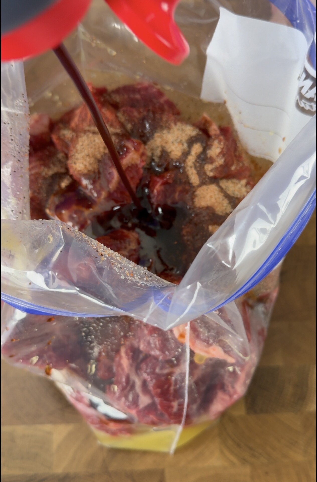 Soy sauce is being poured into a plastic bag filled with raw meat and spices, likely as part of a marinating process. The bag sits open on a wooden surface.