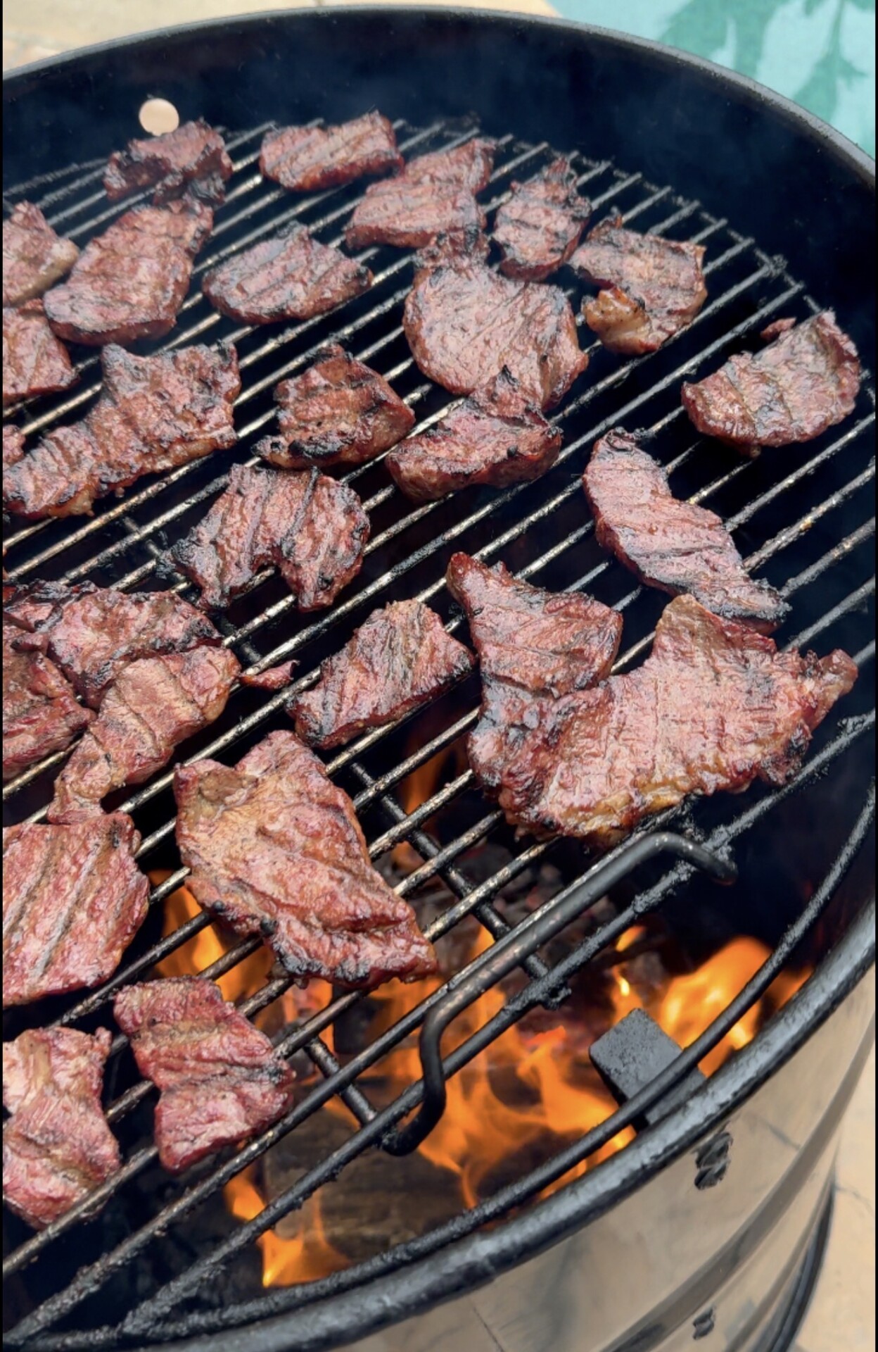 Slices of grilled steak cooking on a round barbecue grill over an open flame, with visible grill marks and flames below.